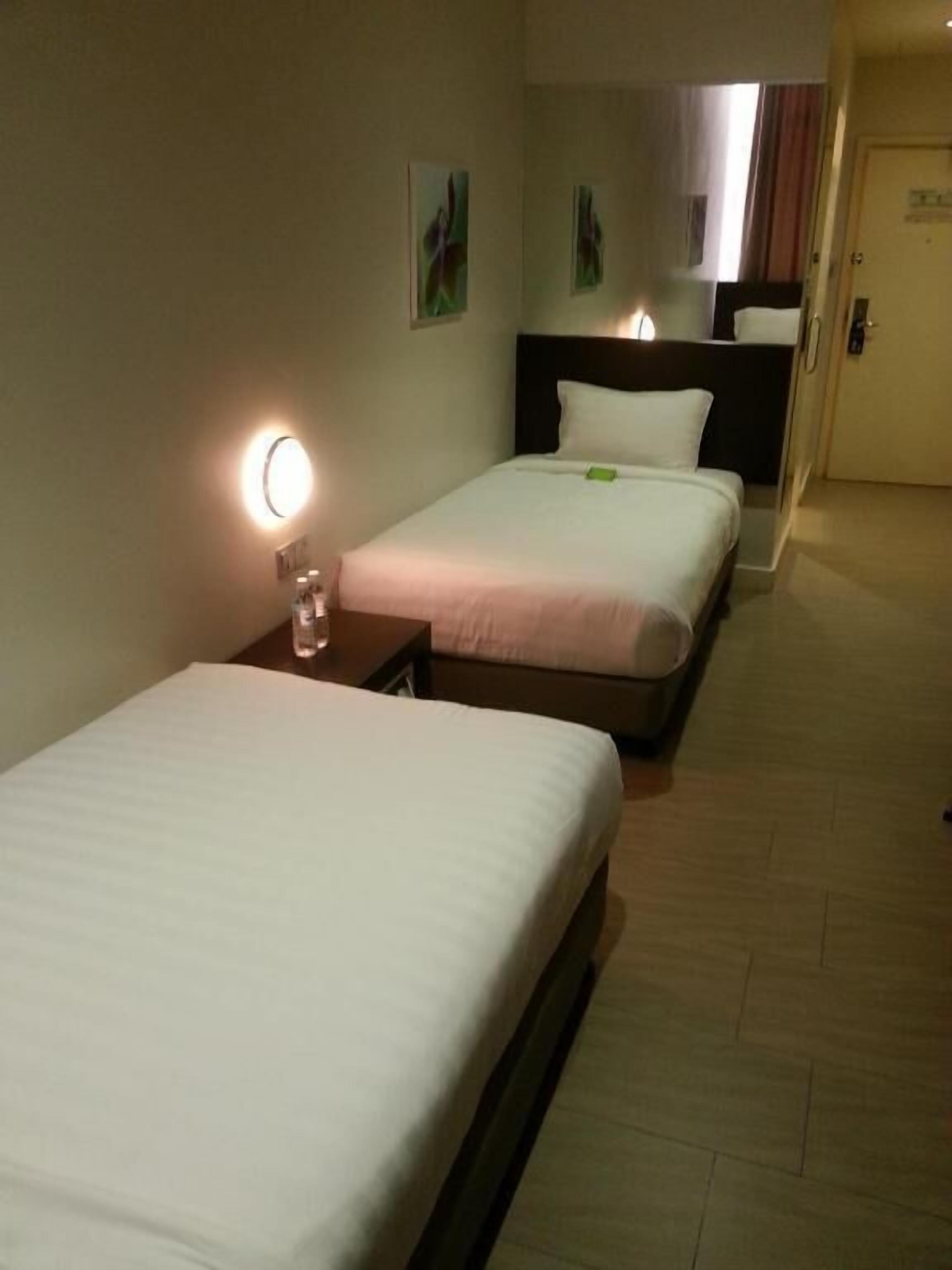 A hotel room with two single beds, a bedside table with a lamp, and a door leading to another room.