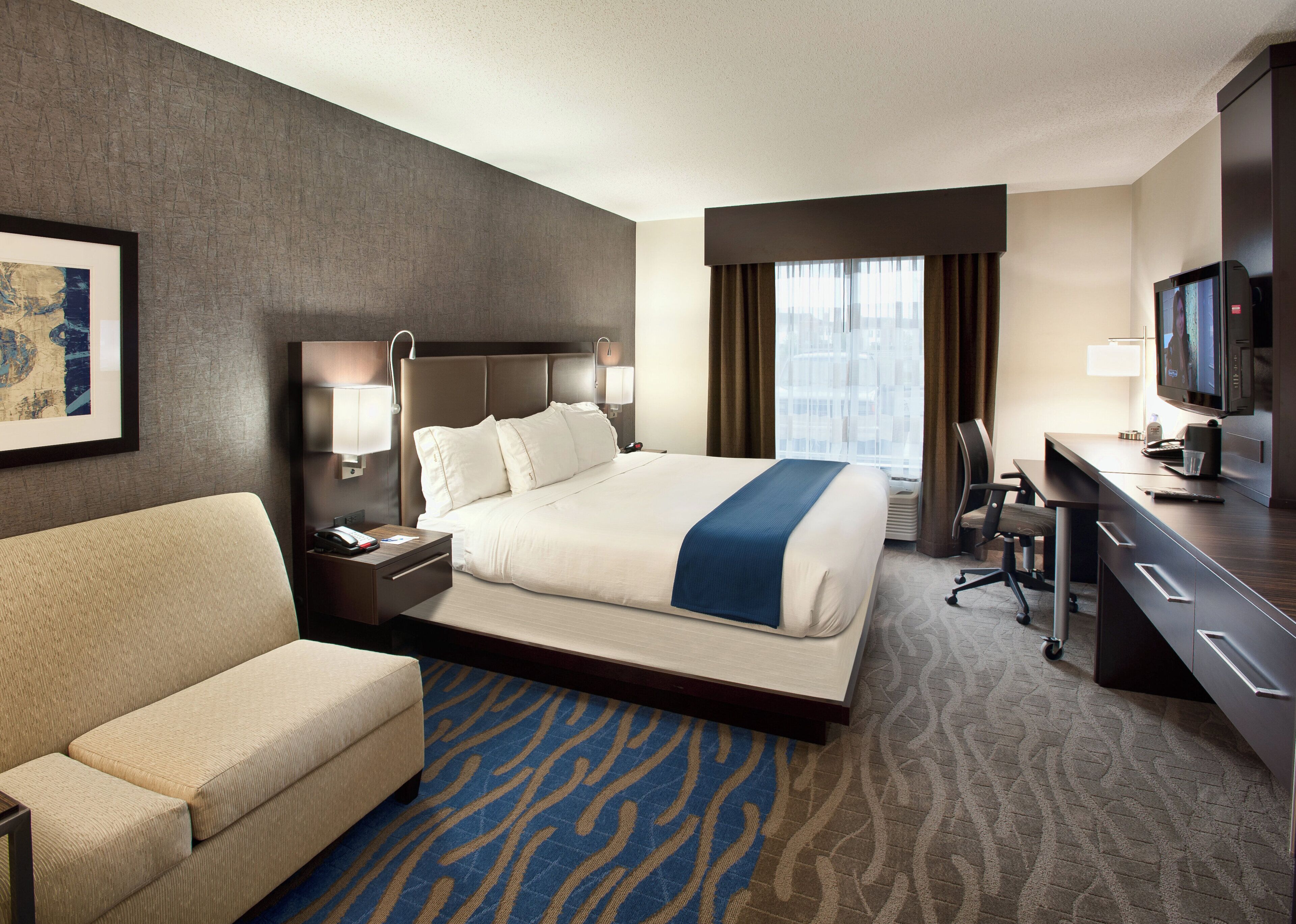 A hotel room with a bed, a desk, a chair, a sofa, a TV, and a framed picture on the wall.
