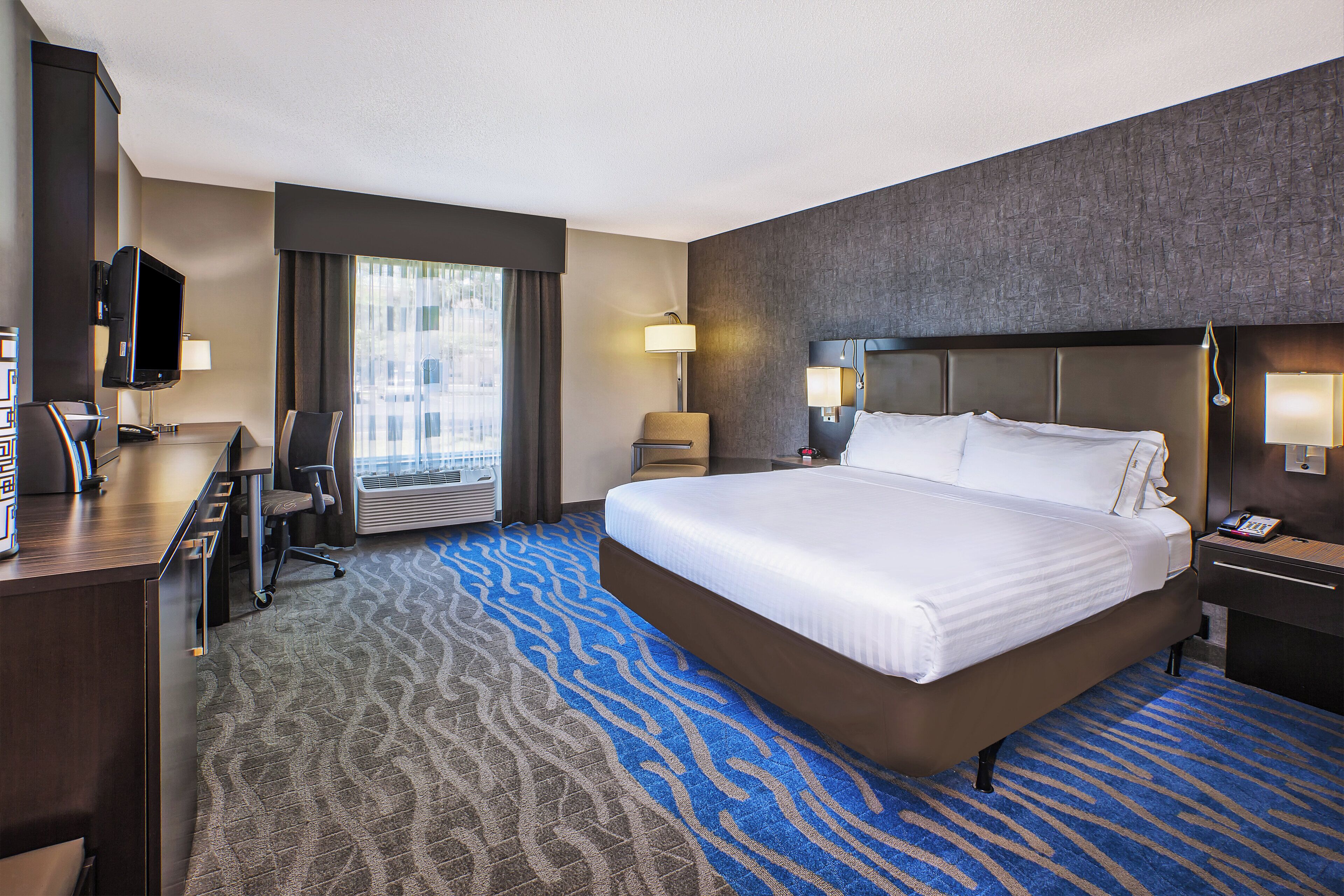 A hotel room with a large bed, a desk with a chair, a television, and a window with curtains.