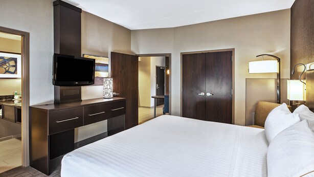 Een hotelkamer met een groot bed, een flatscreen-tv aan een kastje, een bureau met lades, een lamp en een deur die naar een andere kamer leidt.