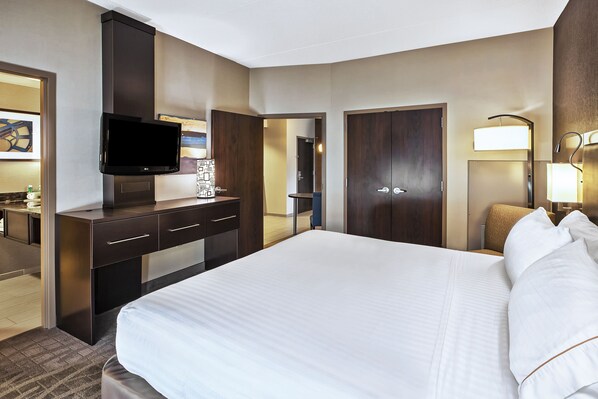 Habitación de hotel con una cama grande, un televisor de pantalla plana montado en un mueble, un escritorio con cajones, una lámpara y una puerta que da a otra habitación.