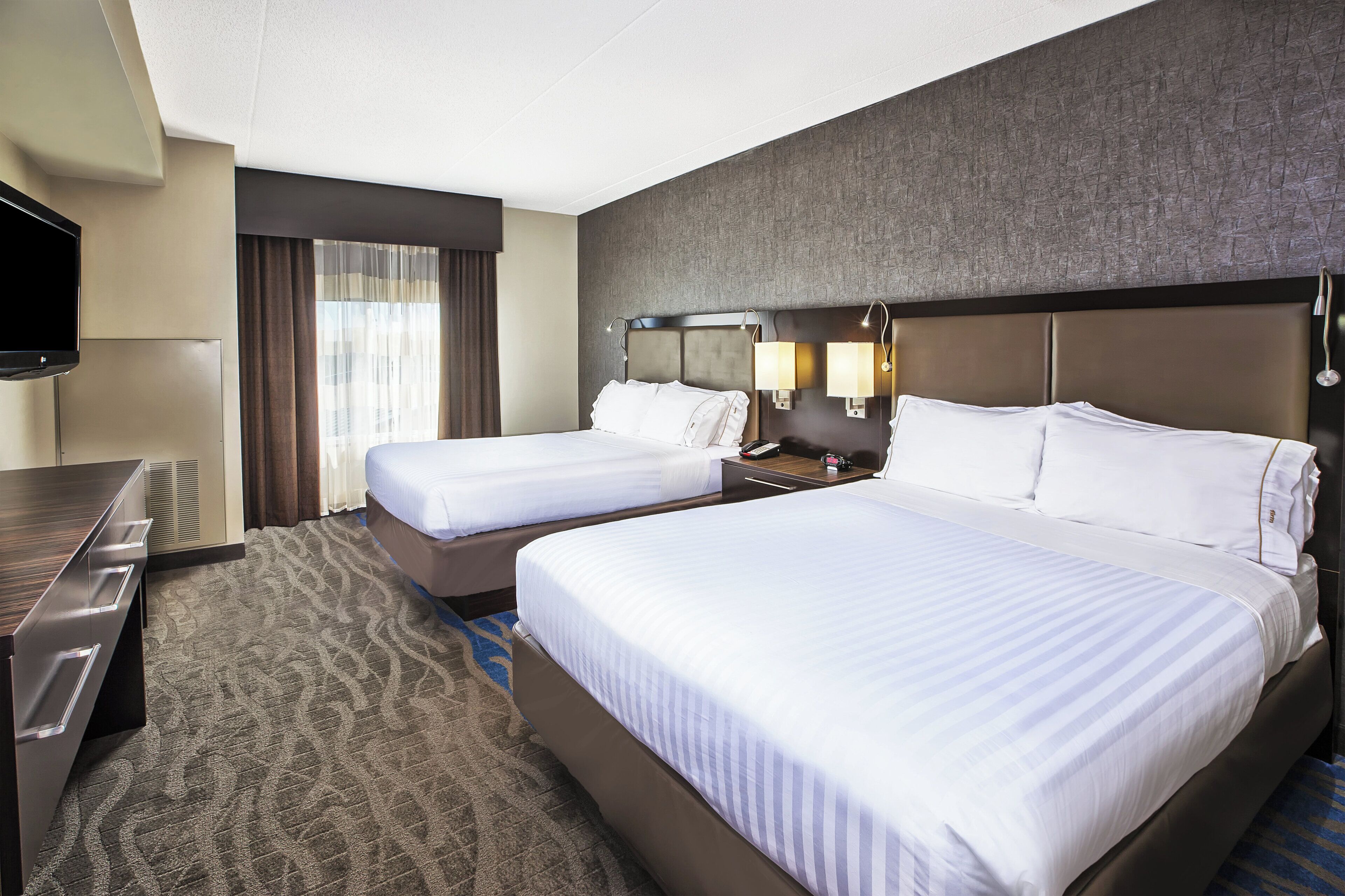 A hotel room with two beds, a desk, a TV, and a window with curtains.