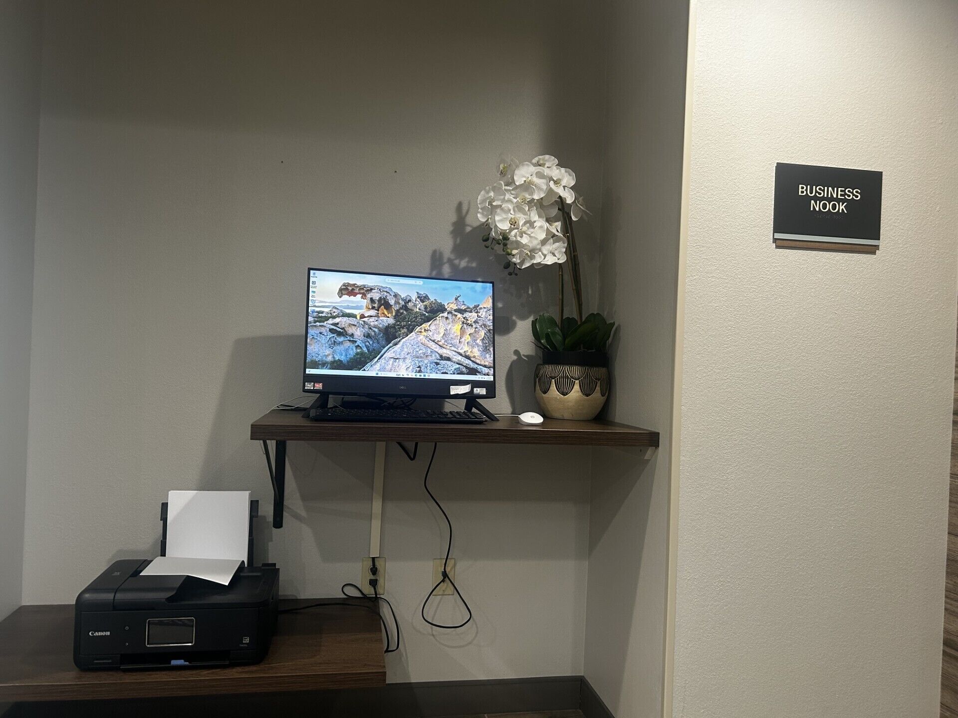 A computer desk with a monitor, keyboard, and mouse, a printer, a potted orchid, and a sign reading 'BUSINESS NOOK'.
