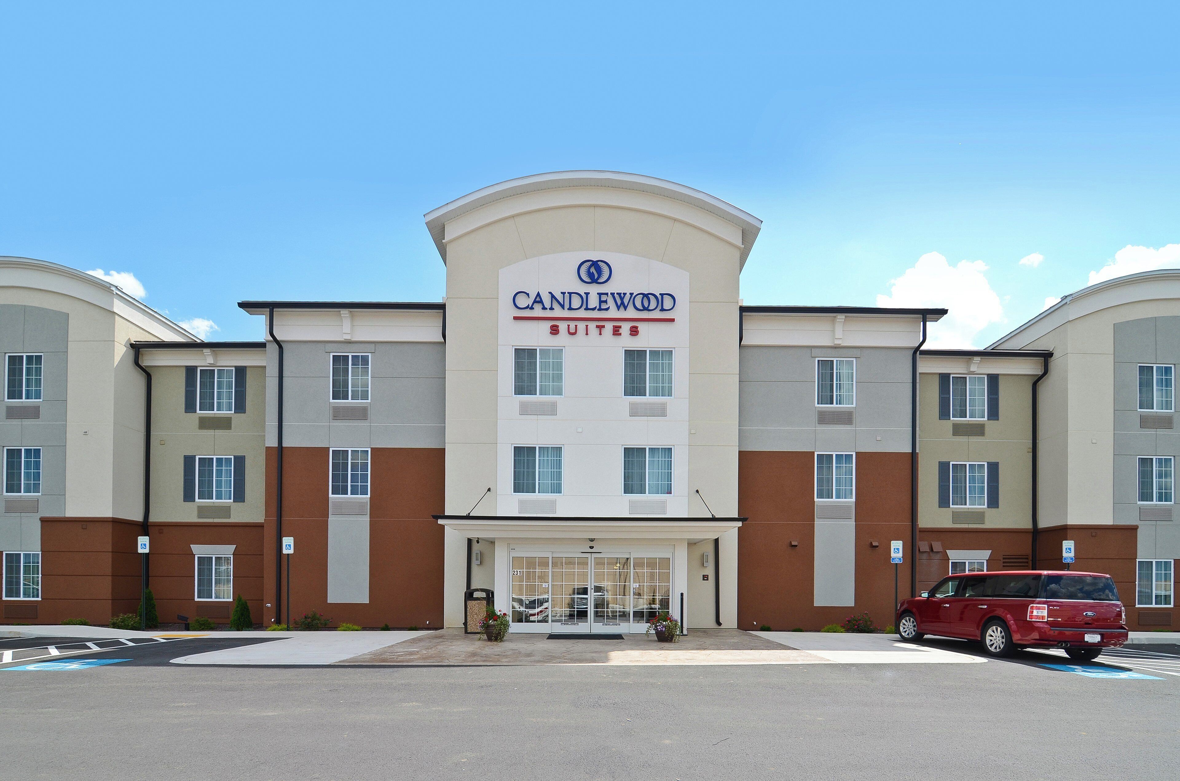 A hotel with a modern facade, a prominent entrance, and a parking area with a red vehicle.