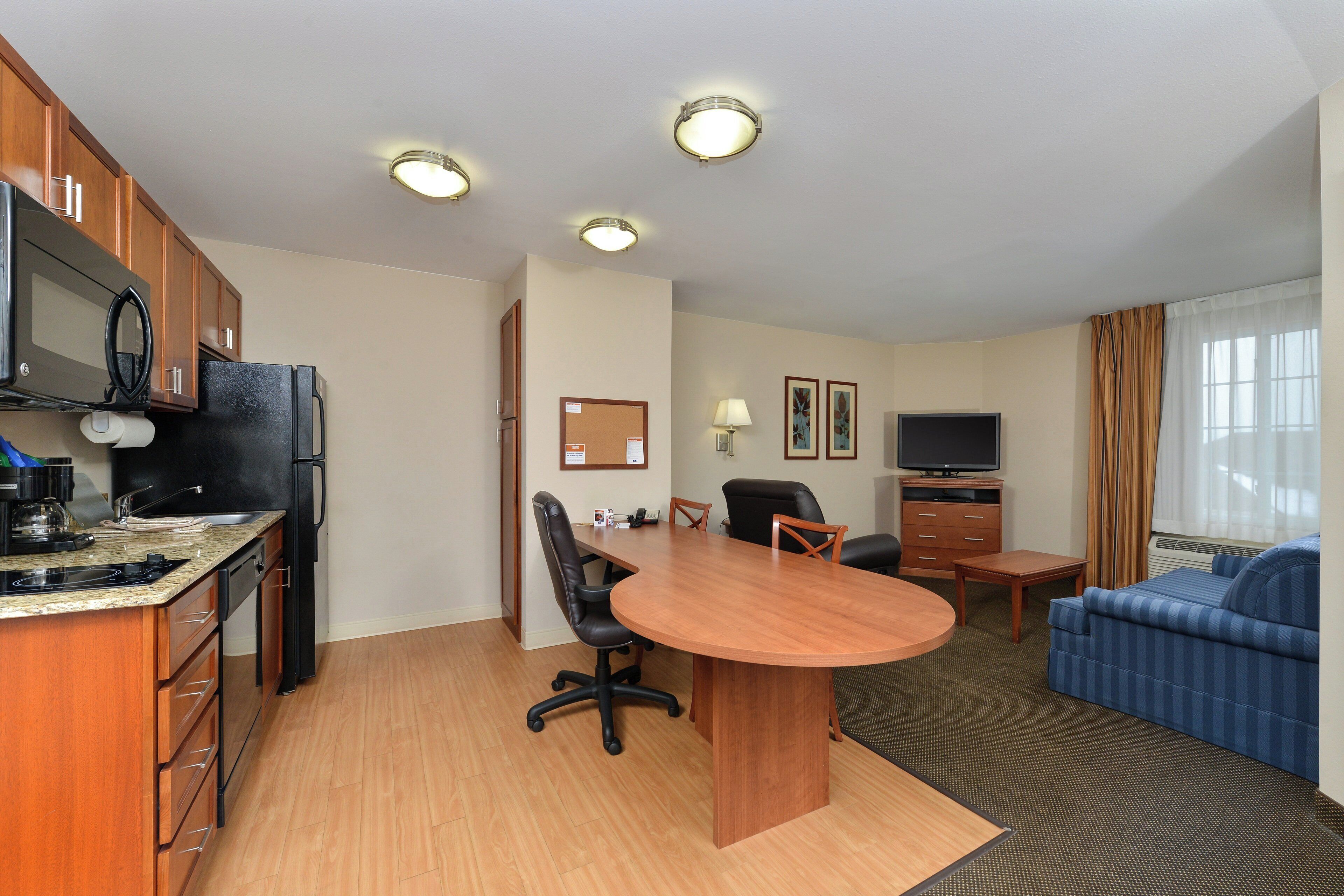 A compact hotel room with a kitchenette, a dining area, a sofa, and a TV.