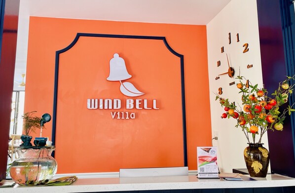 A reception desk with a large sign reading "Wind Bell Villa," a wall clock, a vase with pomegranates, and a teapot on a shelf.