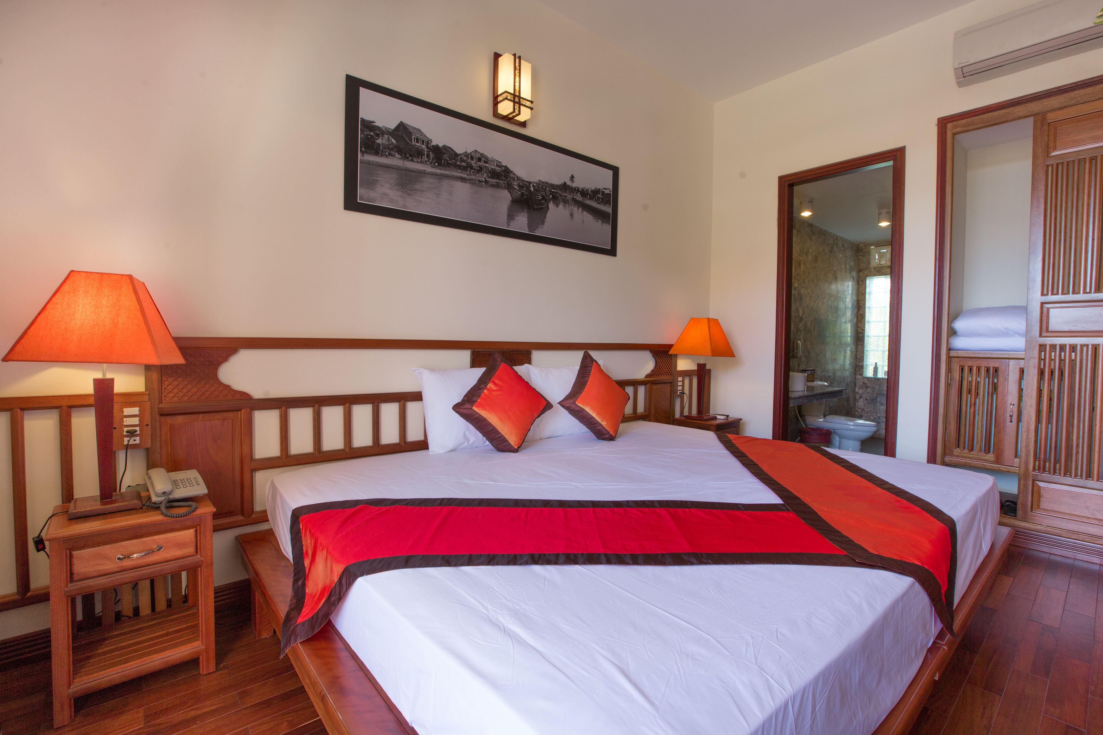 A hotel room with a large bed, two bedside lamps, a nightstand with a phone, a framed picture on the wall, and a wooden wardrobe.