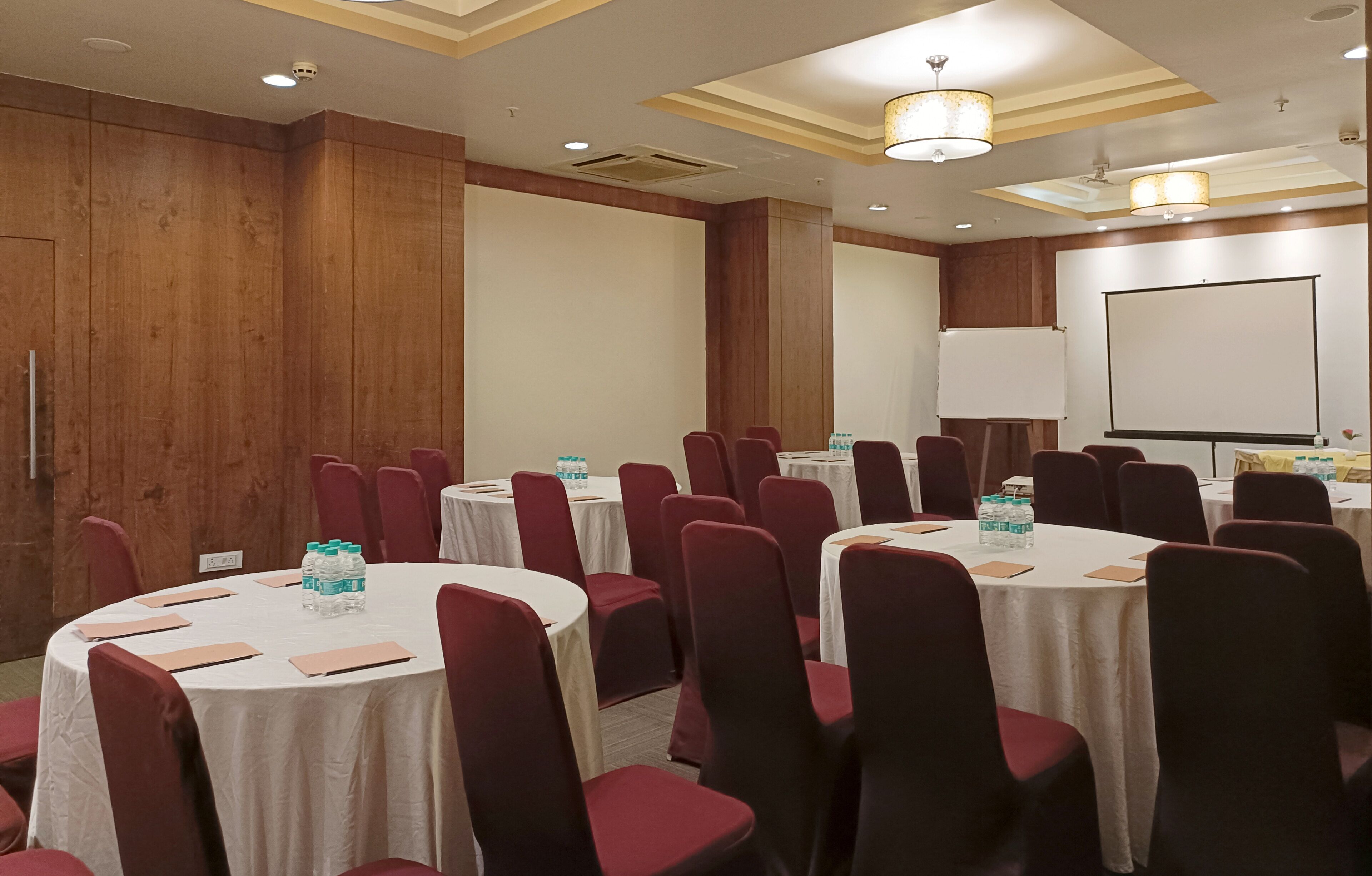 Une salle de conférence équipée de plusieurs tables rondes, chacune entourée de chaises rouges et recouverte de nappes blanches. On peut voir des bouteilles d’eau sur les tables, ainsi qu’un tableau blanc et un écran de projection au fond de la salle.
