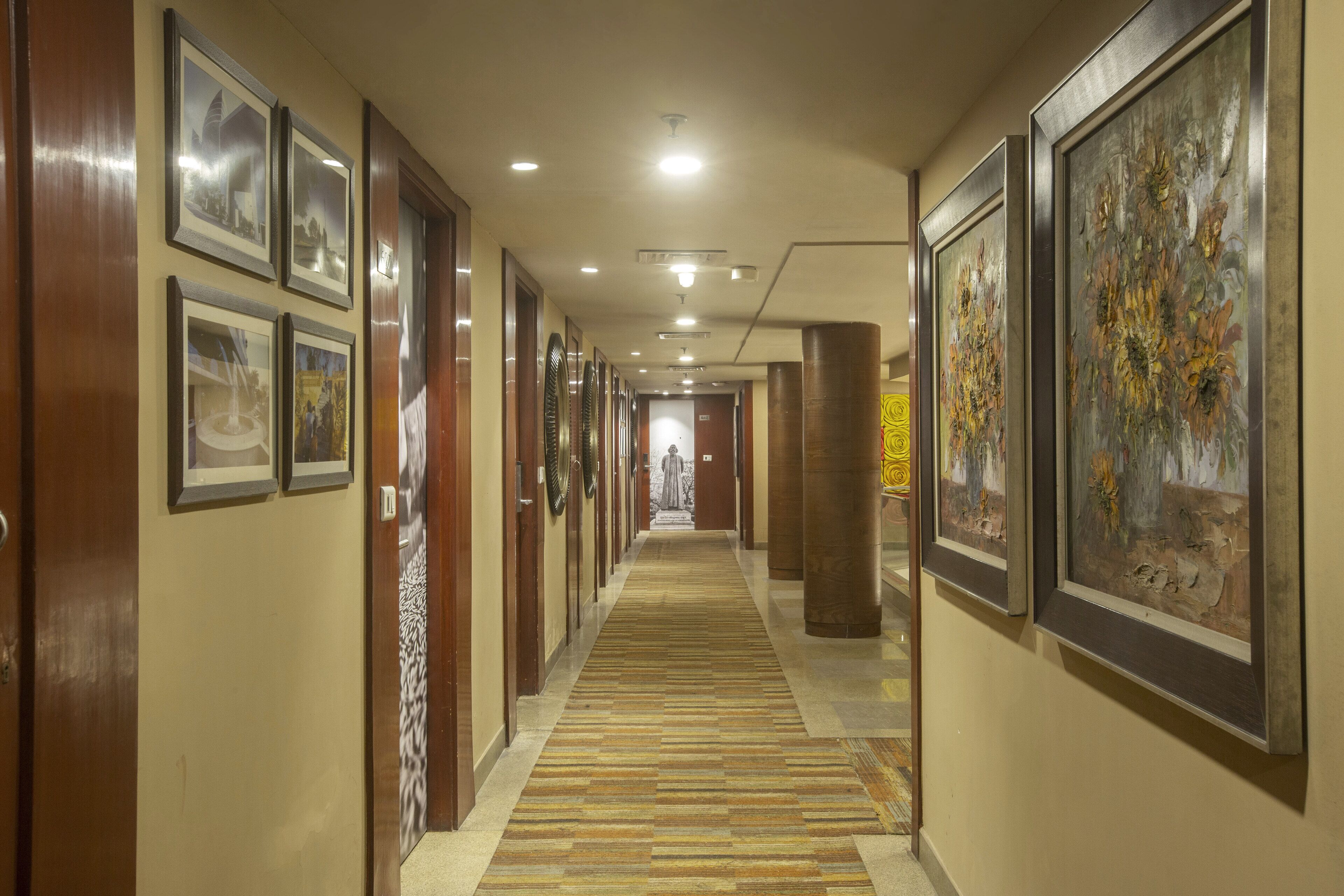 A hotel corridor with framed pictures on the walls, a patterned carpet, and a statue in the distance.