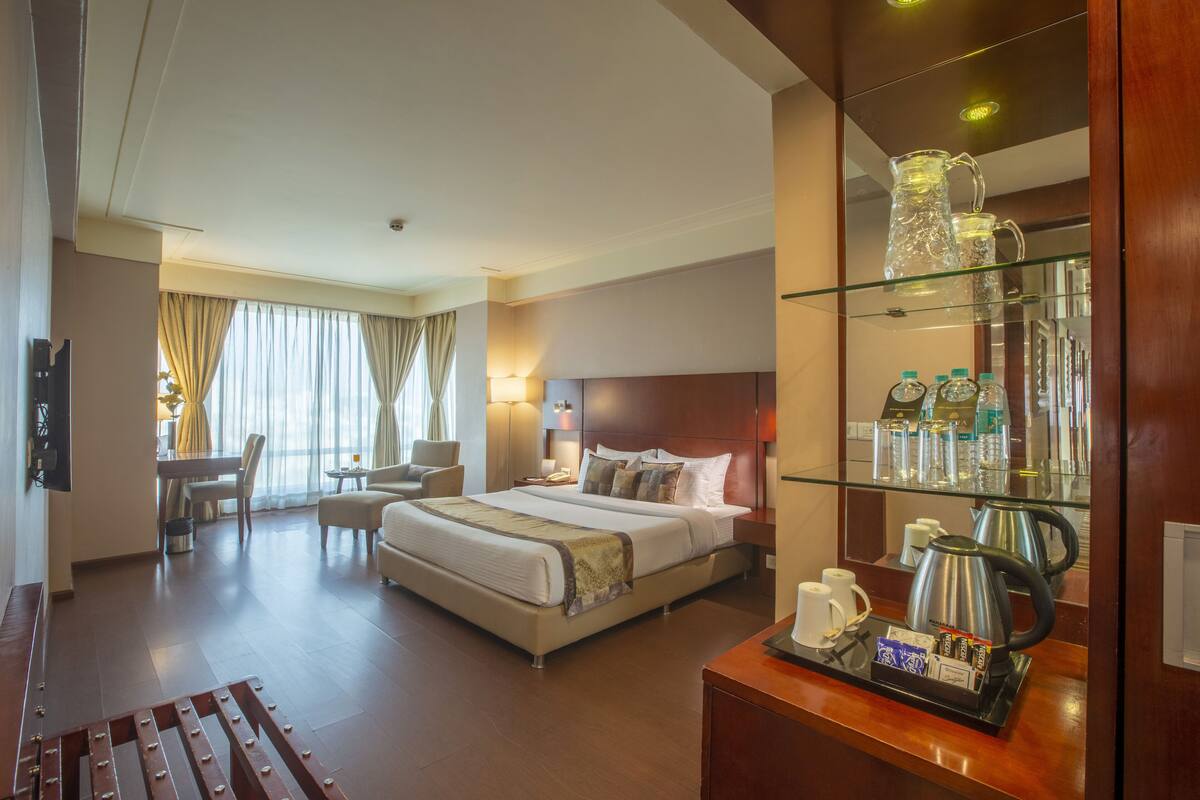 A hotel room with a large bed, a desk with a coffee maker, and a glass display case with bottles.
