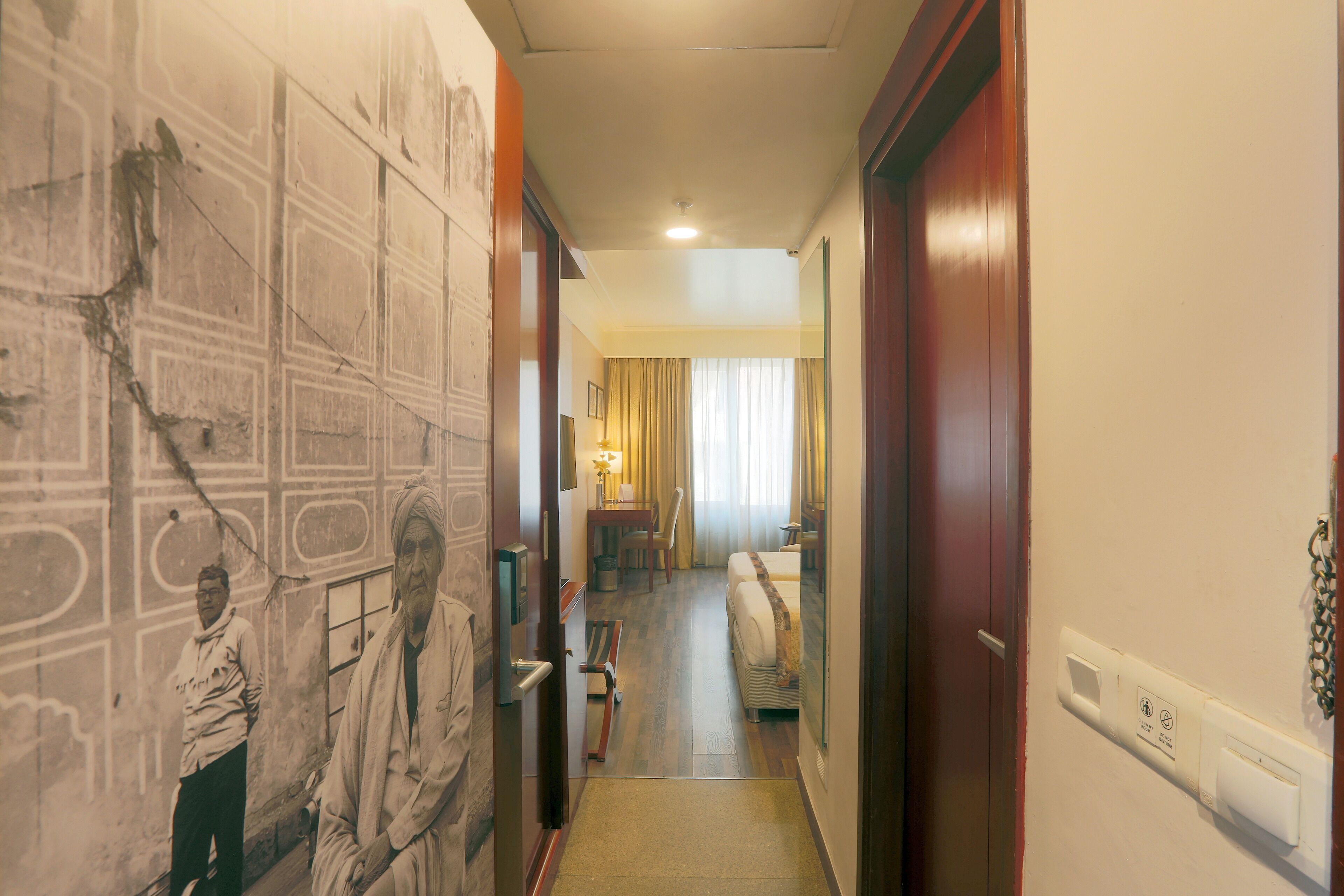 A hotel hallway with a mural, a bed, a desk, and a lamp.