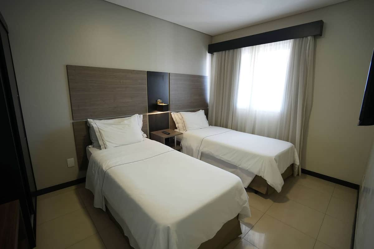 Quarto de hotel com duas camas de solteiro, uma mesa de cabeceira, uma luminária e uma janela com cortinas.