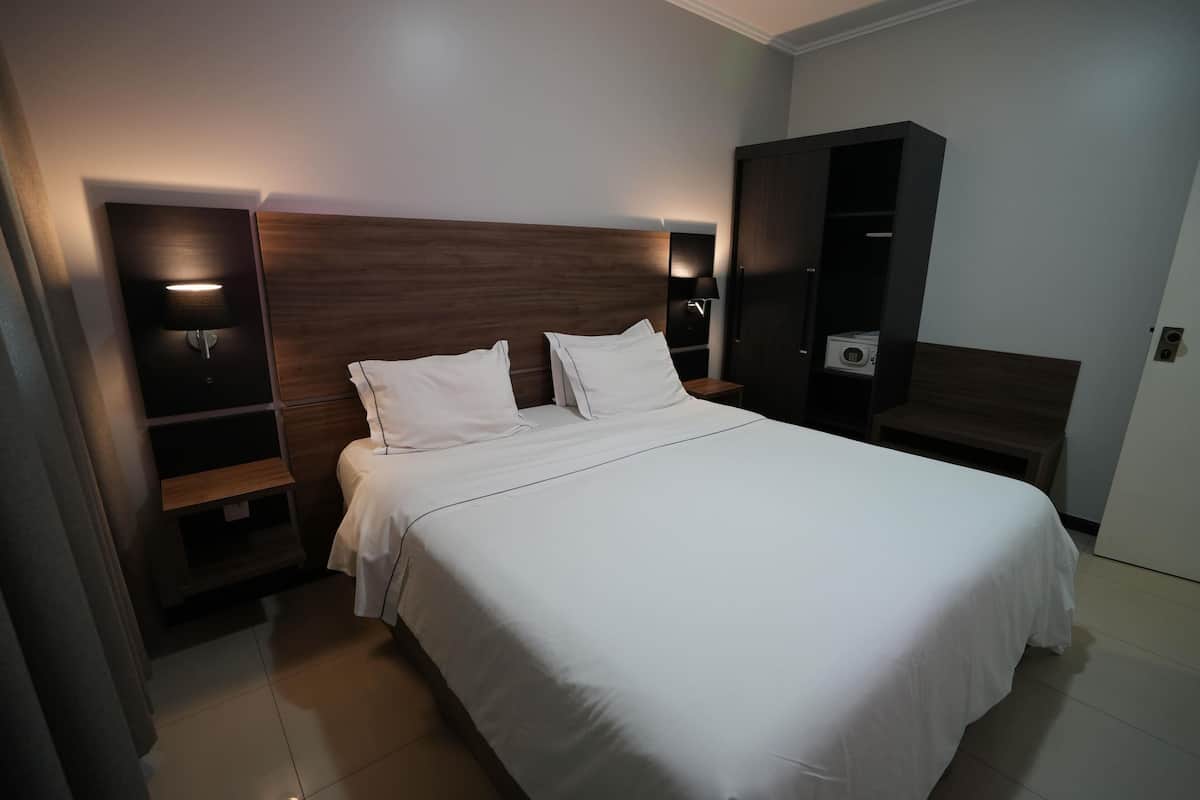 Cama bem arrumada com lençóis brancos, cabeceira de madeira e abajures laterais.