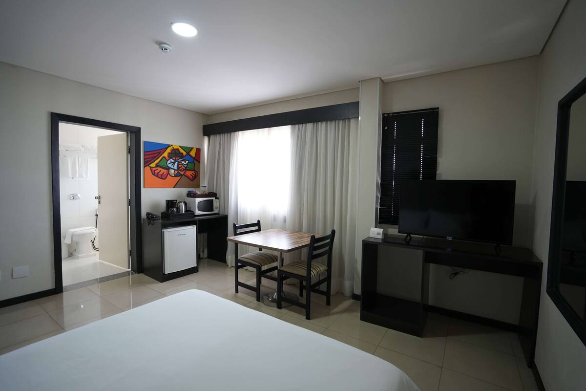 Quarto de hotel com cama, escrivaninha com televisão, micro-ondas, armário pequeno e um quadro na parede.