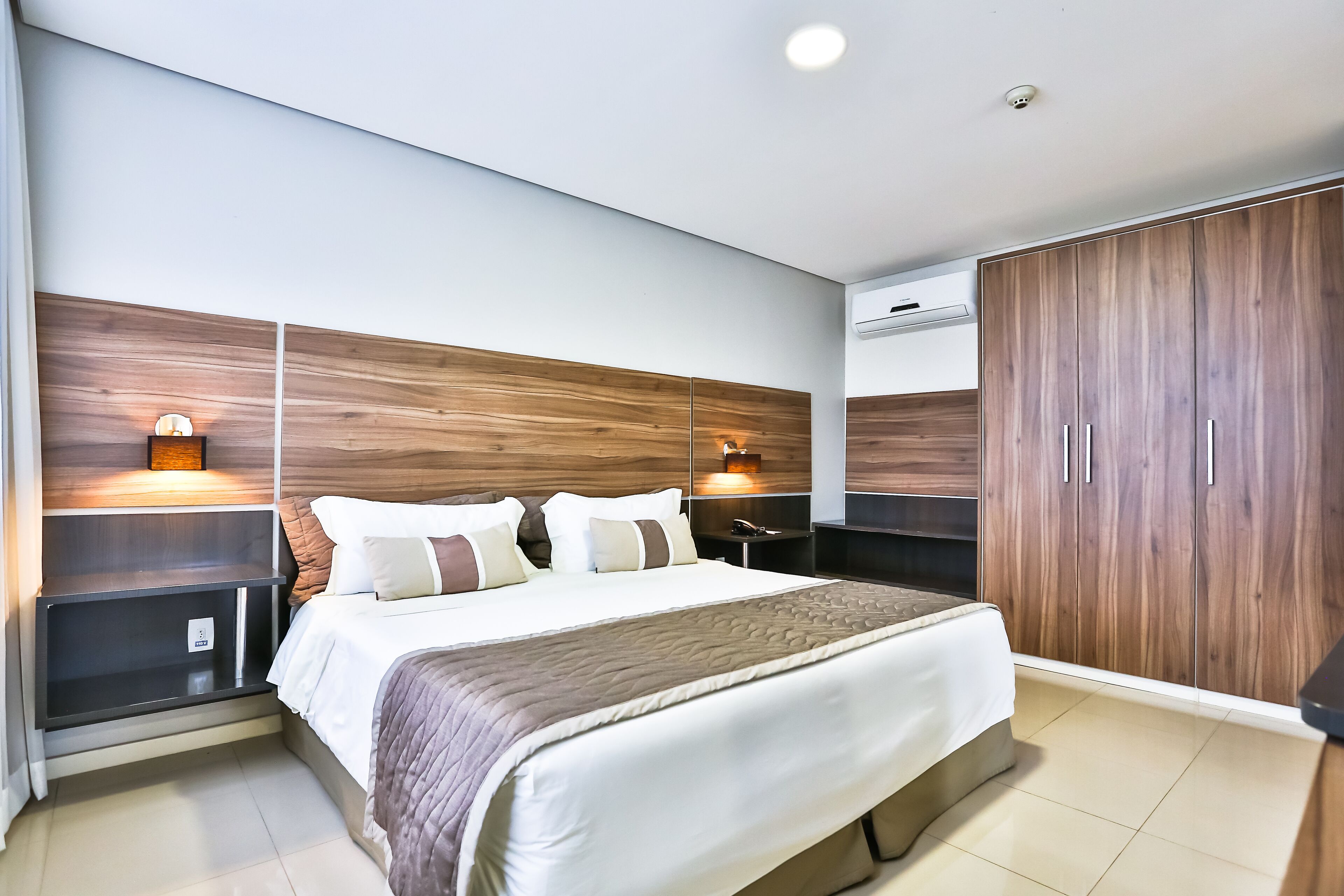 A modern bedroom with a large bed, wooden headboard, bedside lamps, and a wardrobe.