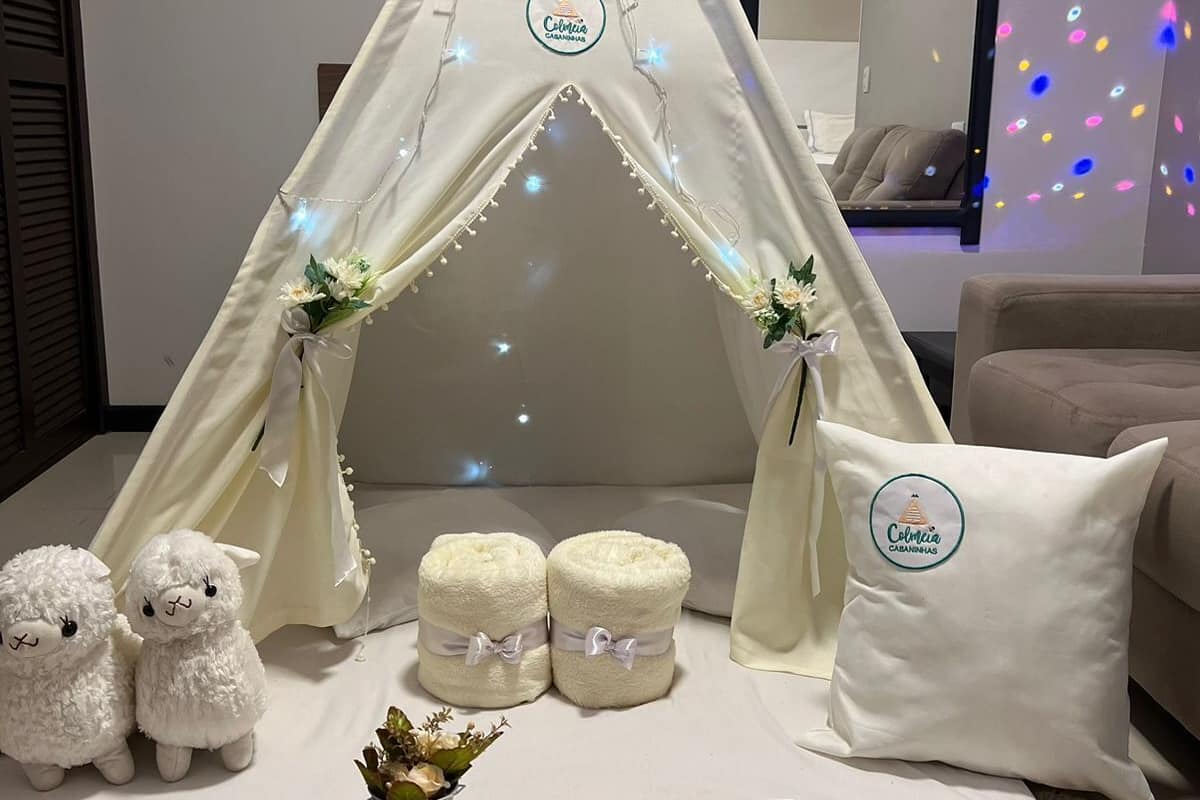 Um quarto aconchegante com uma estrutura branca em forma de tenda, decorada com luzes, dois brinquedos de lã macios e uma pequena planta em vaso.