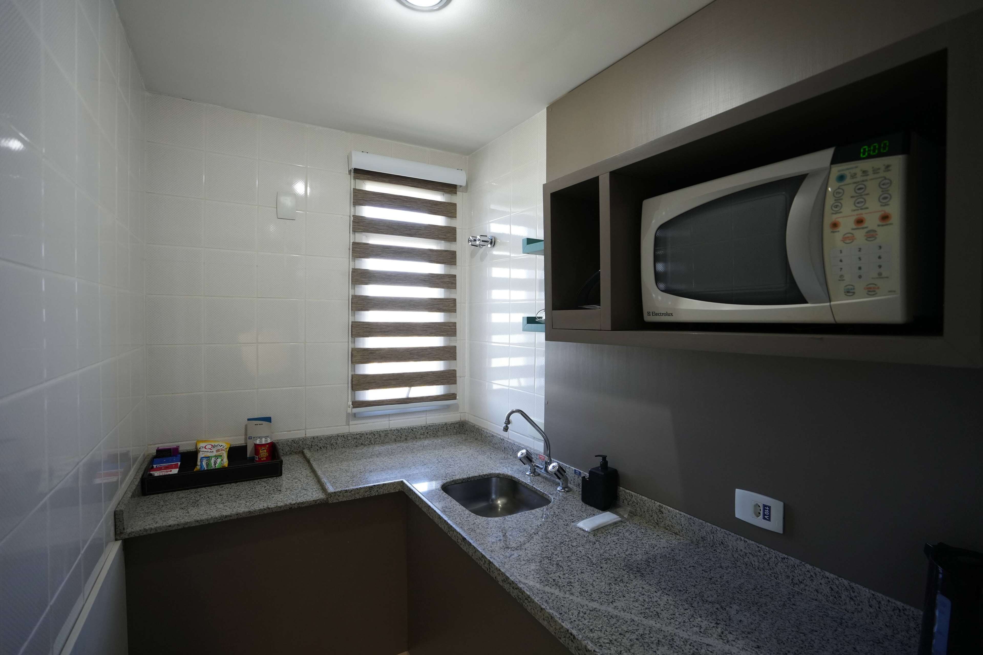 A compact kitchen with white tiled walls, a built-in microwave, a sink, and a window with blinds.