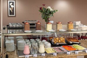 A buffet setup with various food items and beverages, including fruits, pastries, and dairy products, displayed on a glass counter. - Rede Andrade San Martin (Curitiba)