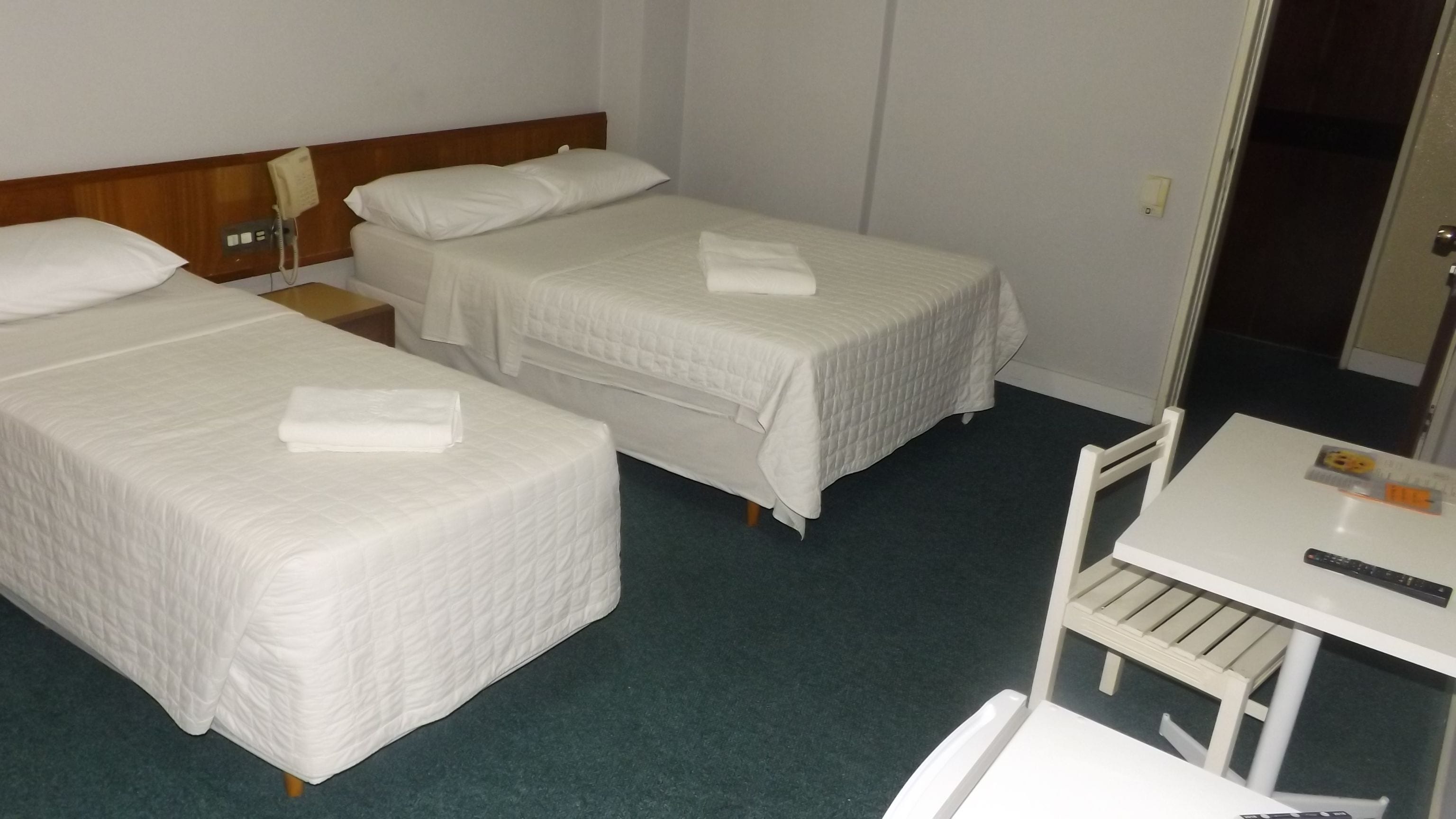 A hotel room with two single beds, a small table with a remote control, and a chair.