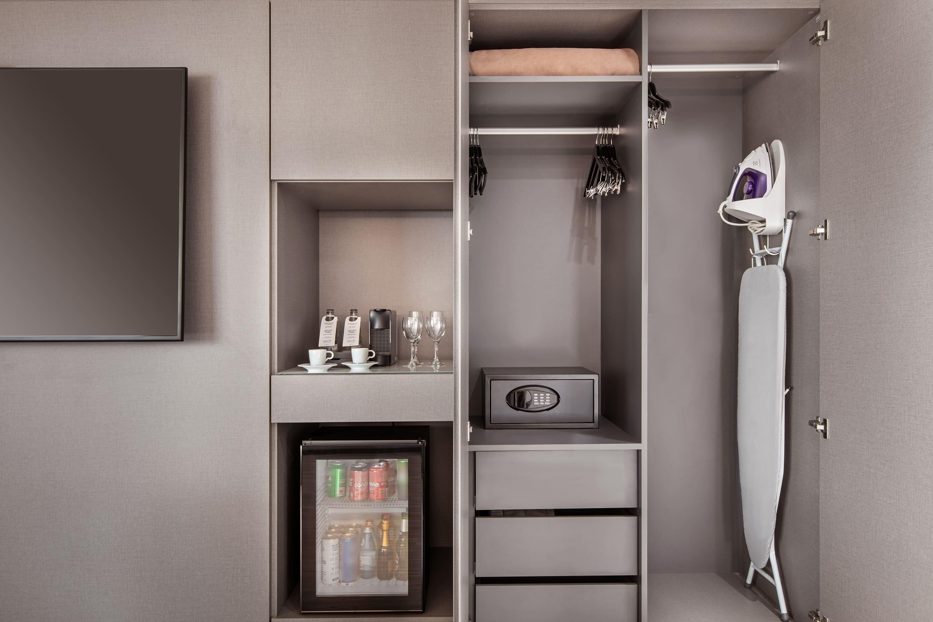 A hotel room with a minibar, a refrigerator, a safe, a hanging iron, and a coat rack.