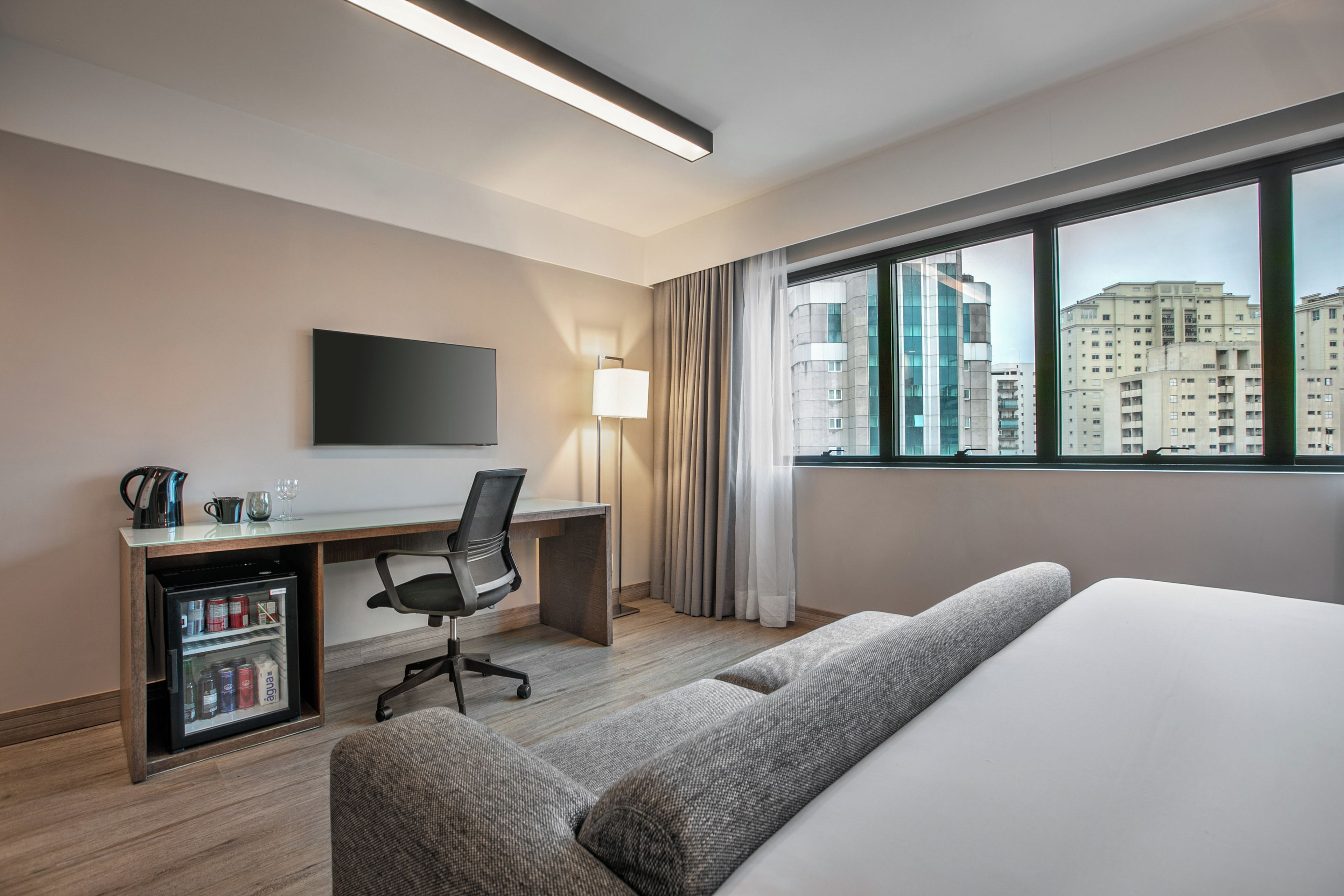 Quarto de hotel com cama, escrivaninha, cadeira, televisão e minibar.