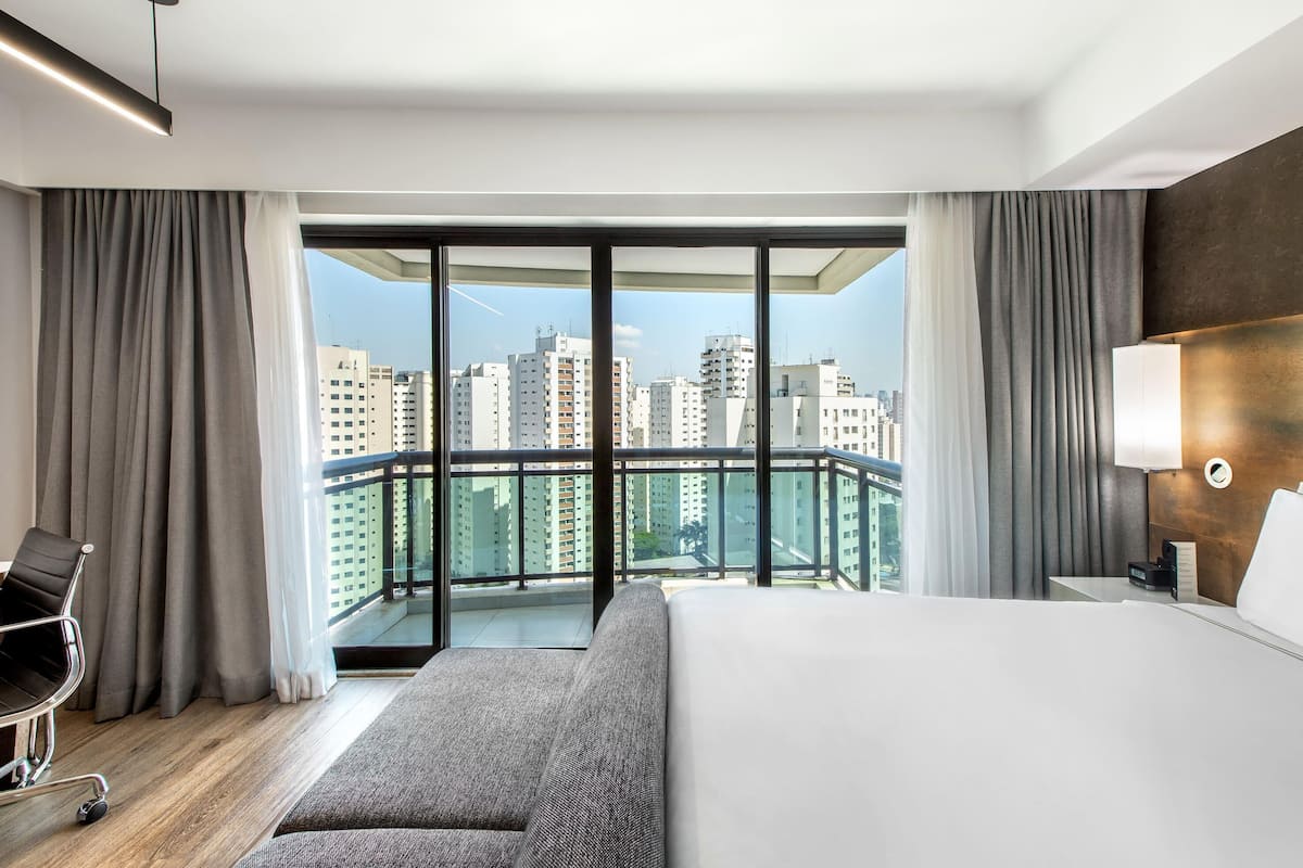 Quarto de hotel moderno com uma cama grande, um sofá, uma escrivaninha e uma cadeira. O quarto tem uma varanda com vista para a cidade.