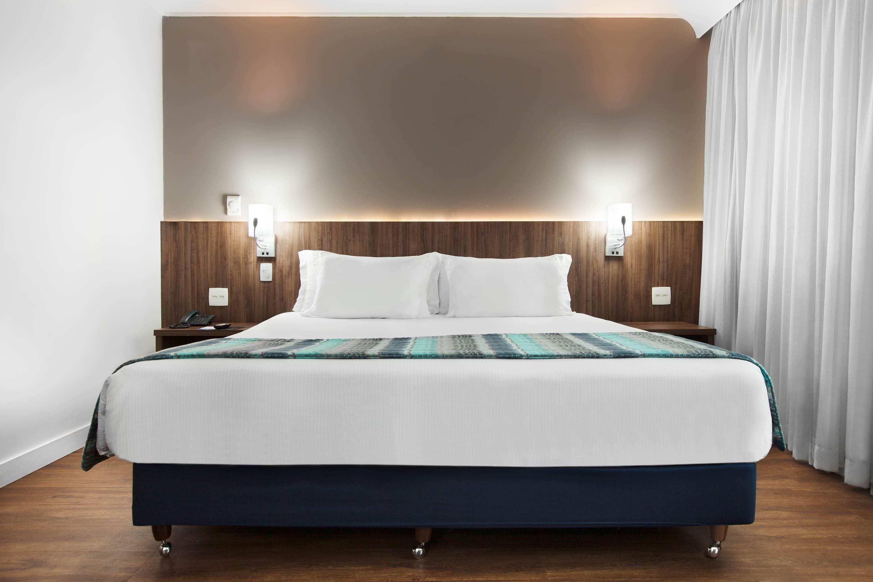 A hotel room with a large bed, wooden headboard, bedside lamps, and a curtain on the window.