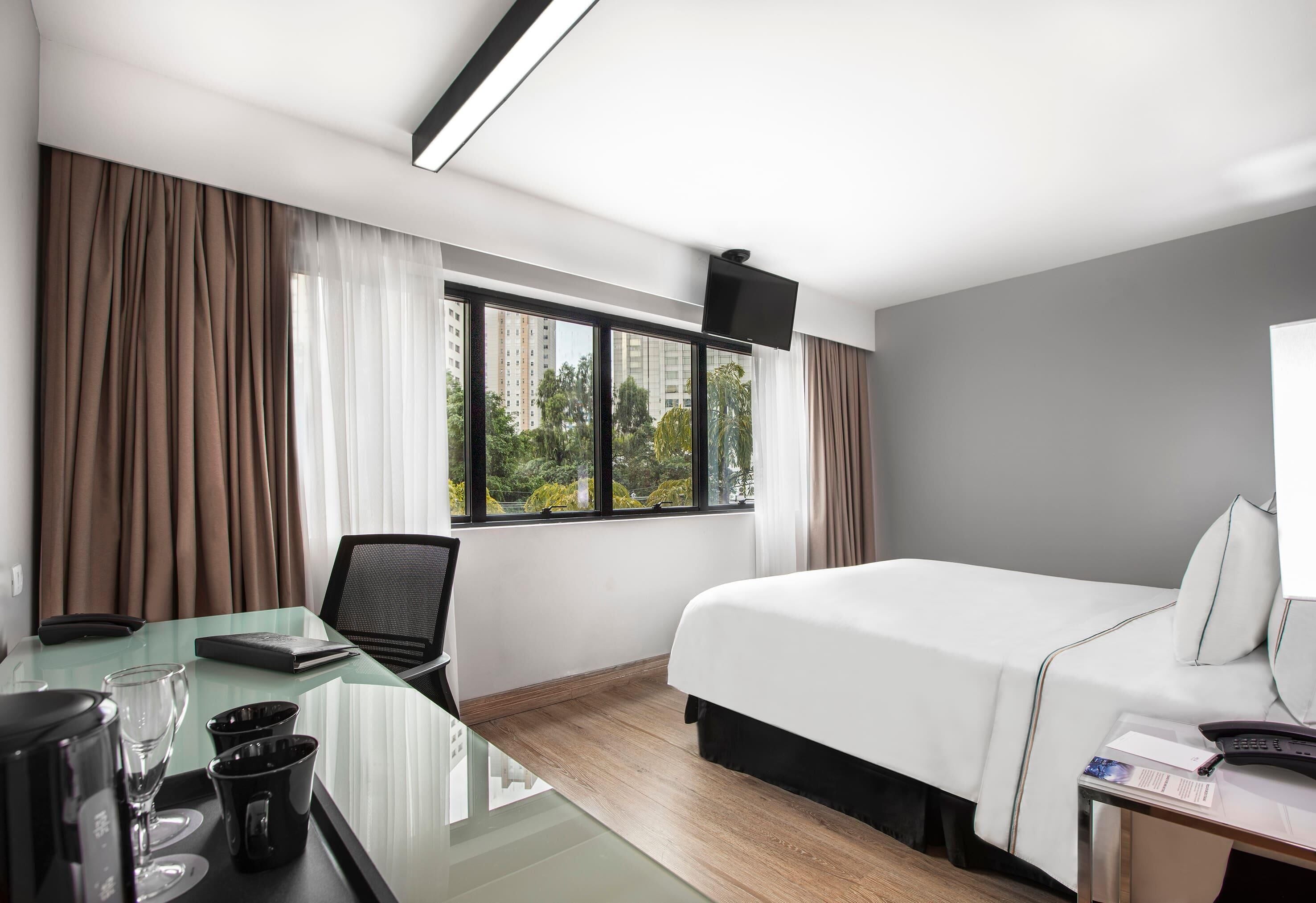 Una habitación de hotel con una cama, un escritorio con una computadora portátil, una silla, una cafetera y una ventana con cortinas.