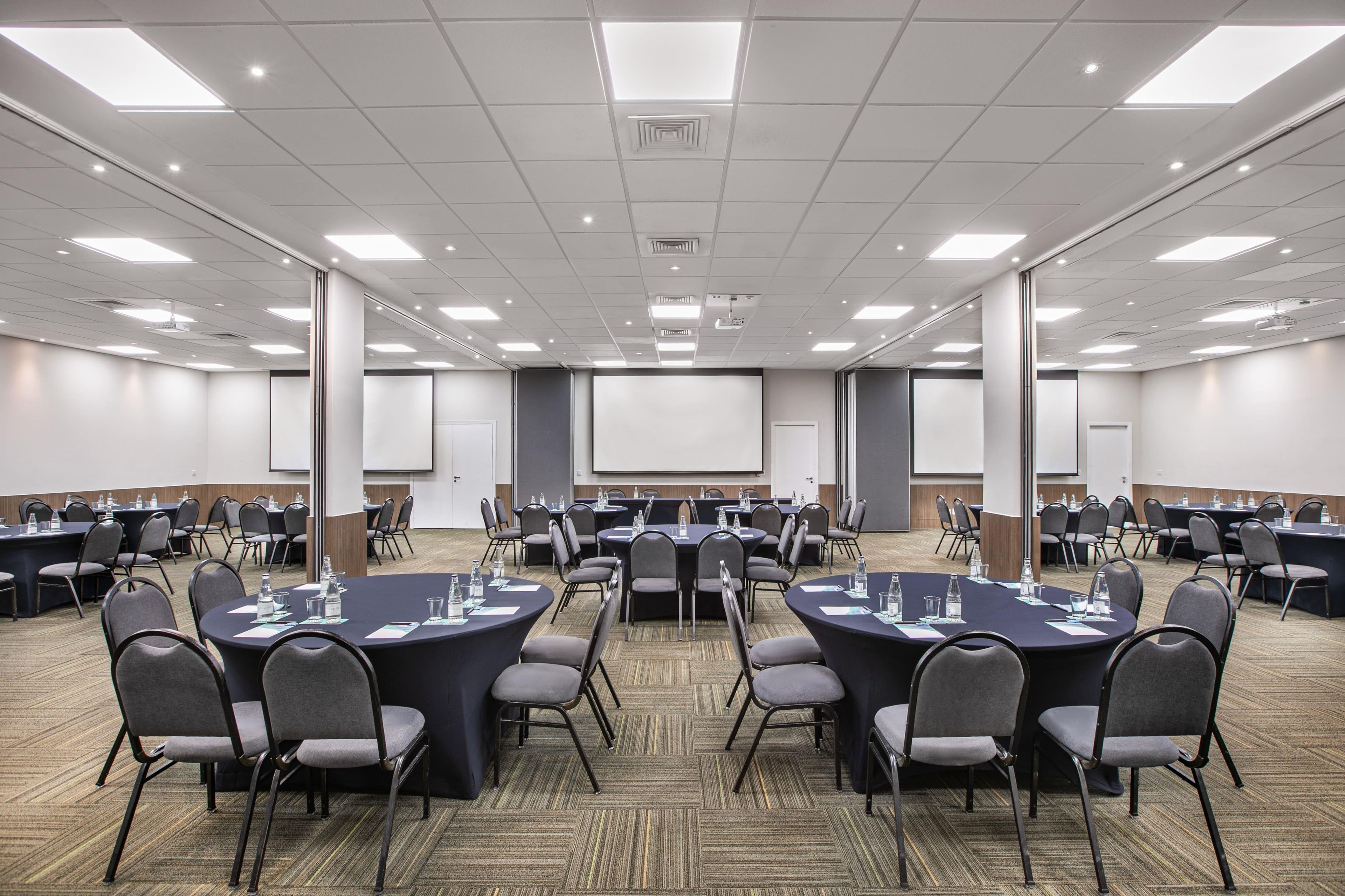 A conference room with multiple round tables, chairs, and a large screen at the front.