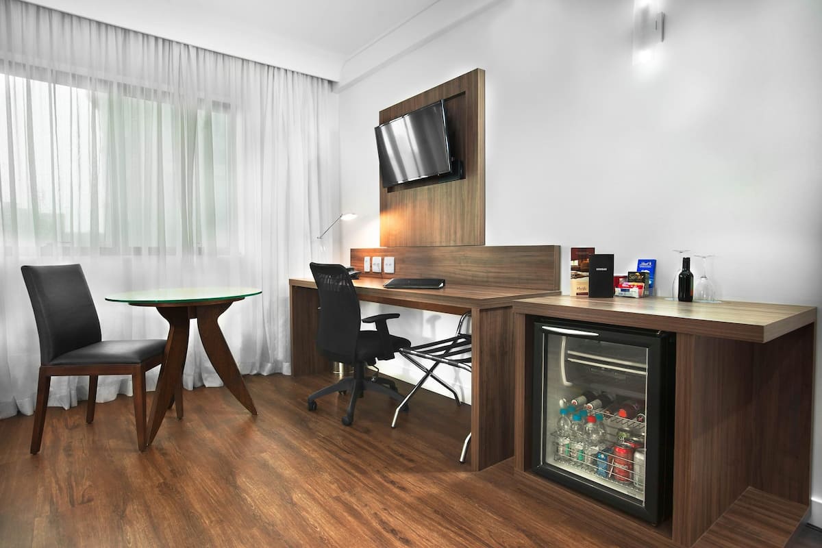 Quarto de hotel com escrivaninha de madeira, uma cadeira, televisão fixada na parede e mini-geladeira com bebidas.