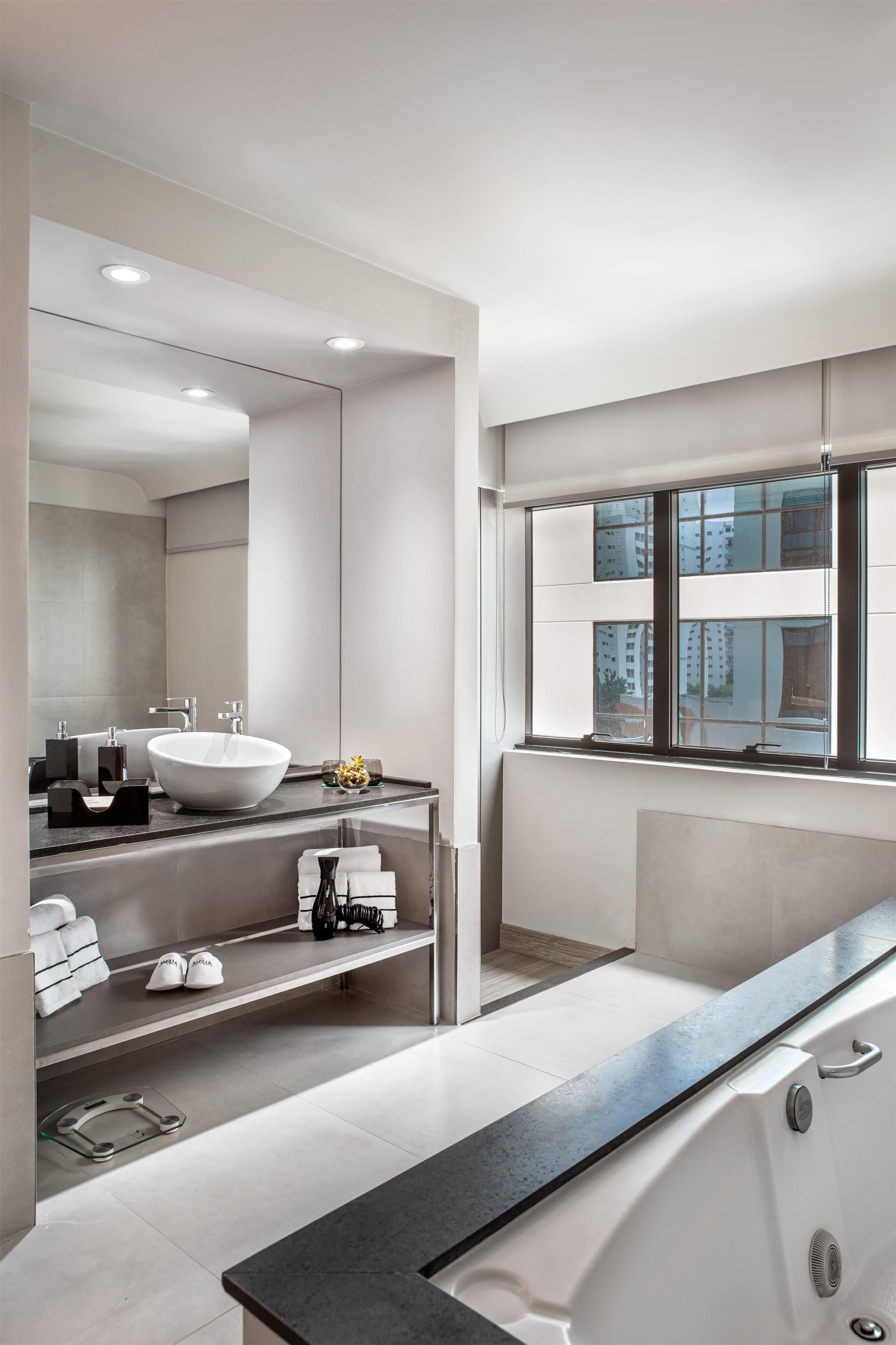 A modern bathroom with a large mirror, a white sink, and a bathtub. There is a window with a view of buildings outside.