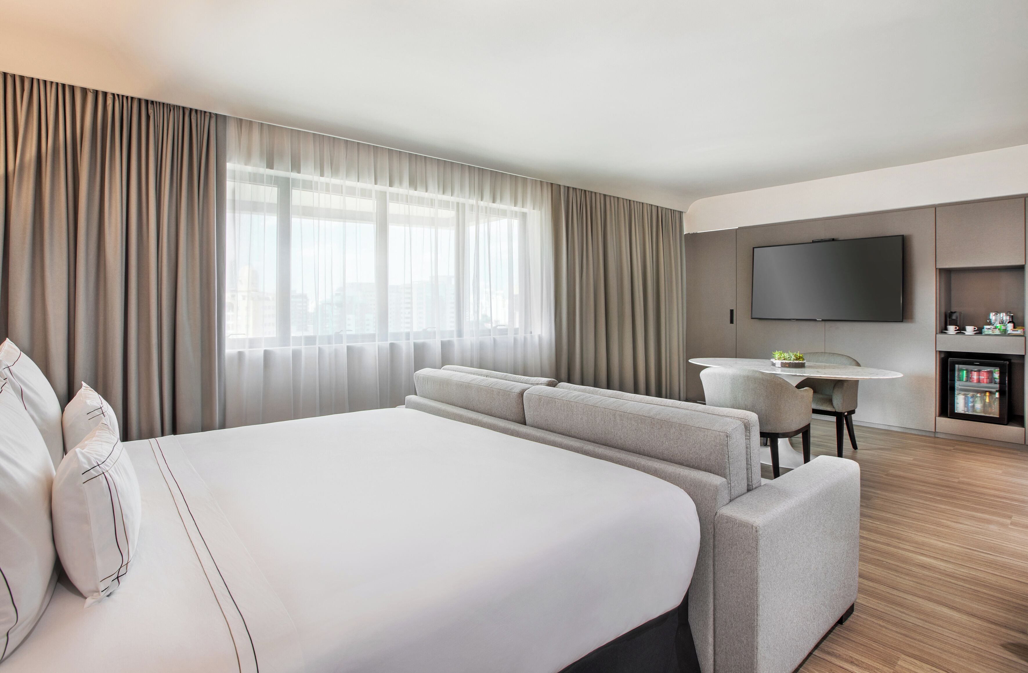 A hotel room with a large bed, a seating area with a table, a television, and a minibar.