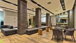 Un área de descanso moderna con sofá, mesas redondas y sillones, que presenta suelo de madera y pilares de piedra.