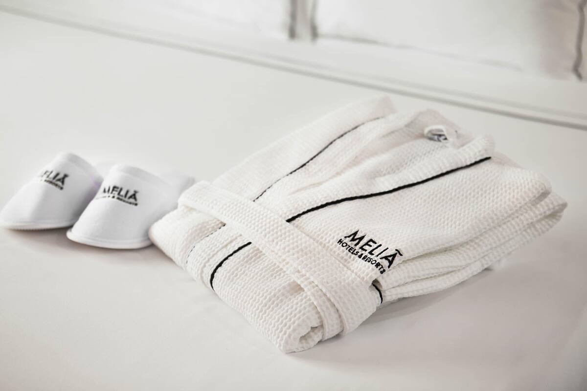 Uma toalha branca dobrada, com a marca 'MELIA', e um par de chinelos sobre uma superfície branca.