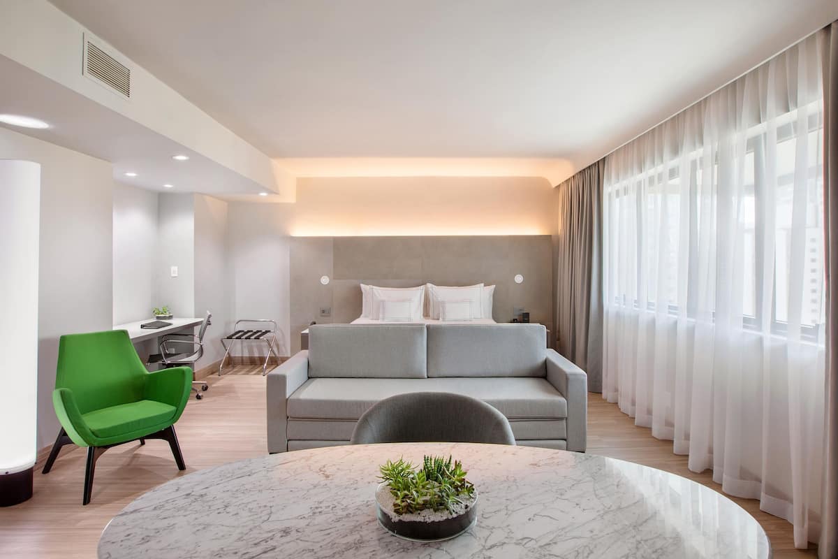 Quarto de hotel moderno com sofá cinzento, poltrona verde, mesa redonda de mármore com uma planta e cortinas leves nas janelas.