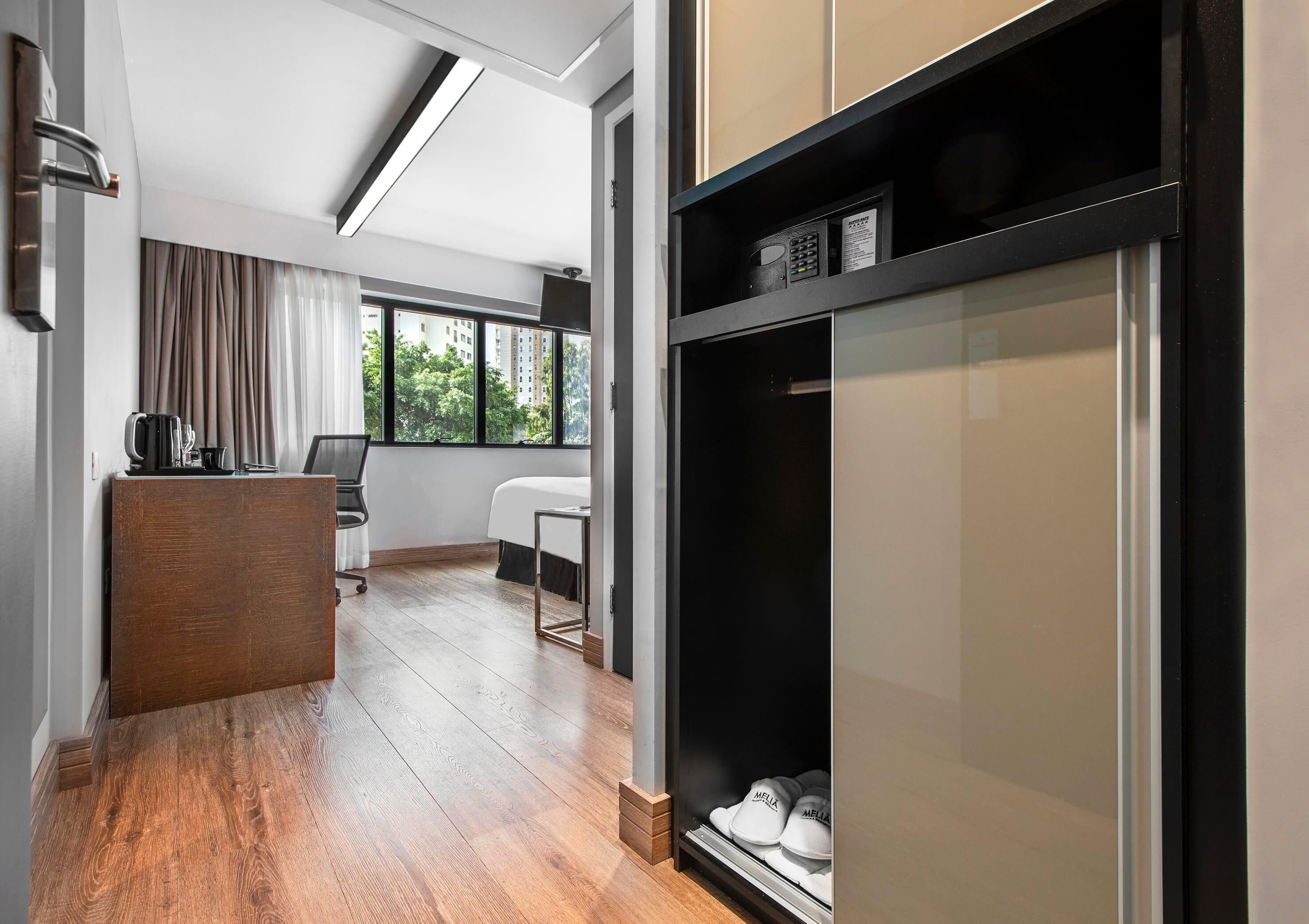 A hotel room with a wooden floor, a desk with a kettle, a chair, a bed, a wardrobe with shoes, and a window with a view of trees.