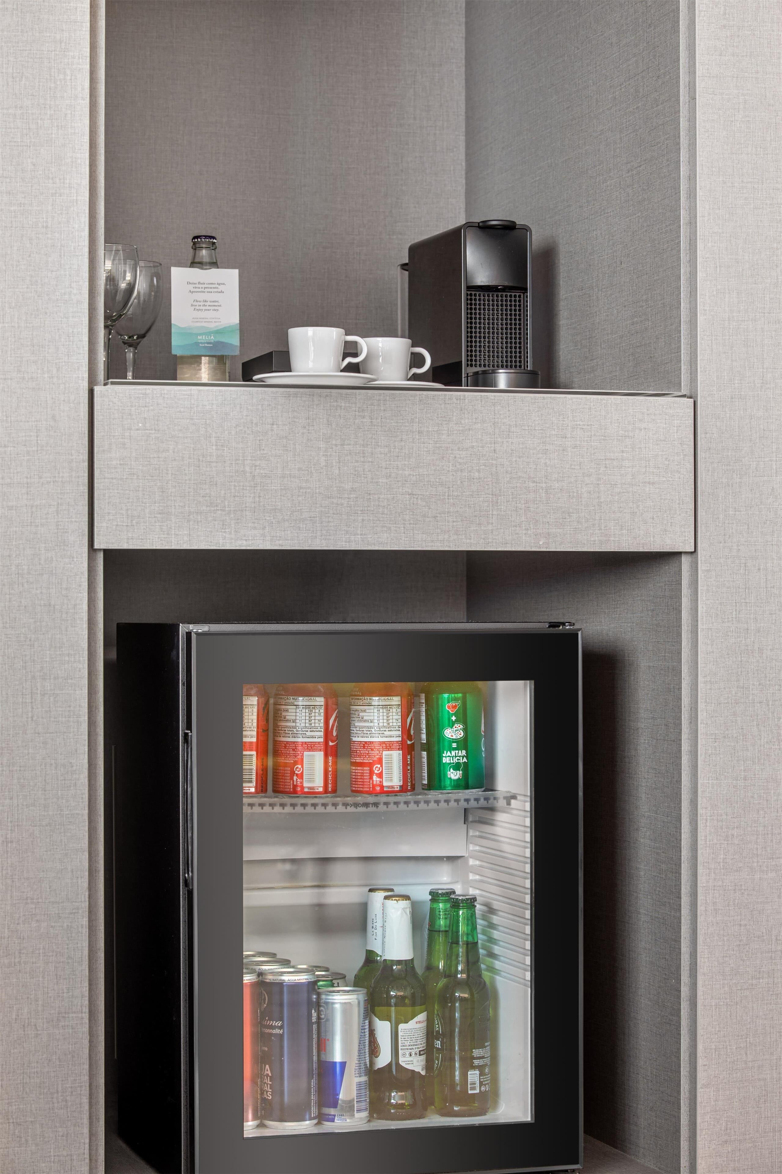 A minibar with a coffee machine, a glass, a bottle, and a cup on the top shelf, and various beverages in the refrigerator below.