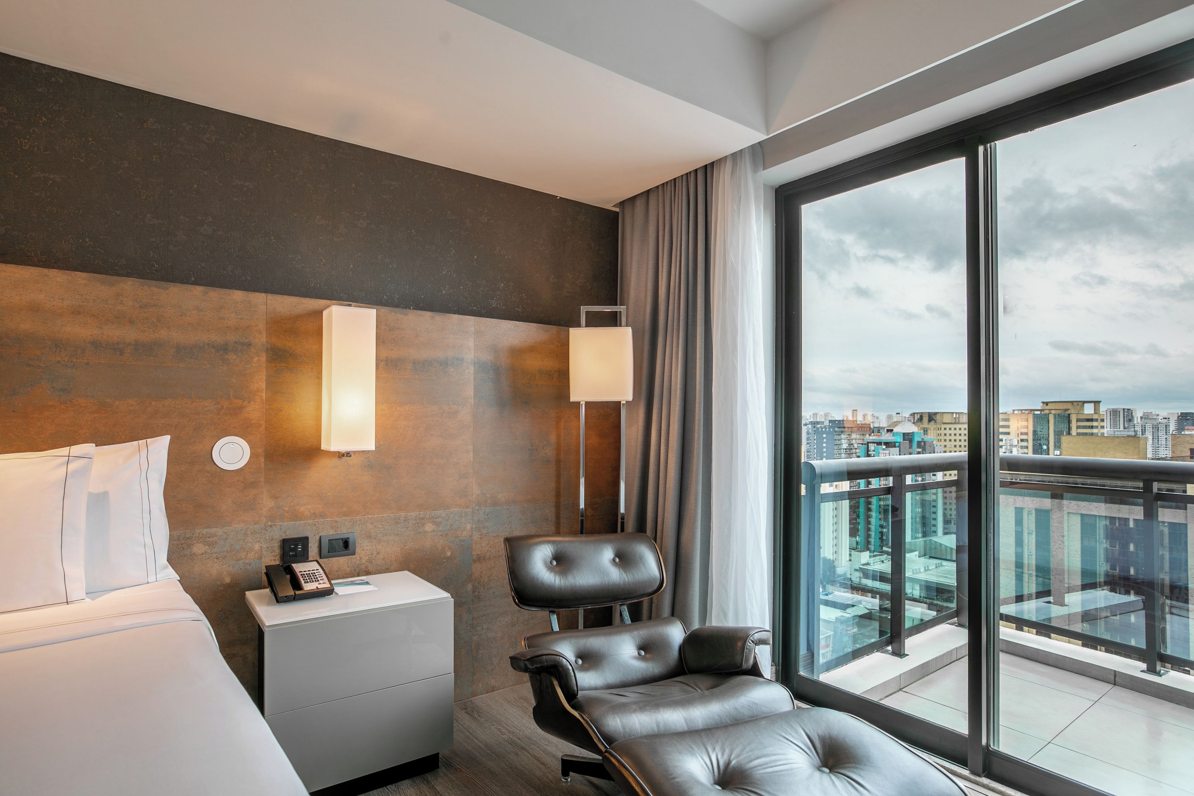 A hotel room with a bed, a desk, a chair, a phone, and a large window with a city view.