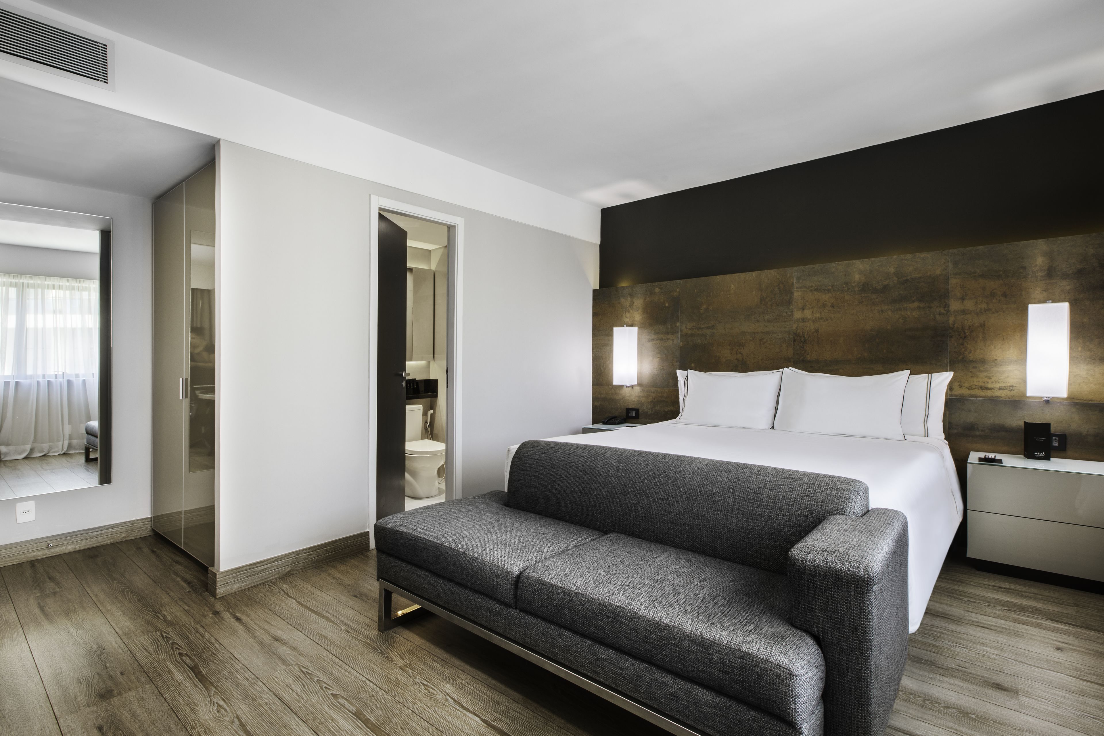 A modern hotel room with a large bed, a sofa, a walk-in closet, and a bathroom visible through an open door.
