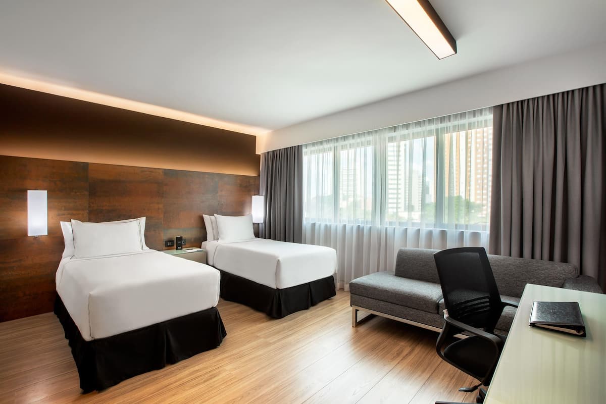 Quarto de hotel com duas camas, um sofá, uma escrivaninha e uma janela grande com cortinas.