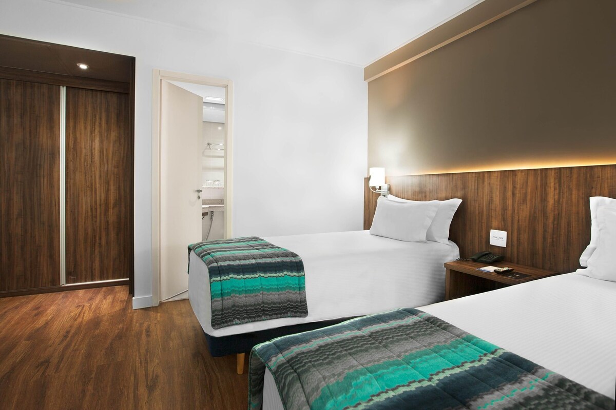 Una habitación de hotel con dos camas, suelo de madera, un armario y una mesita de noche con lámpara.
