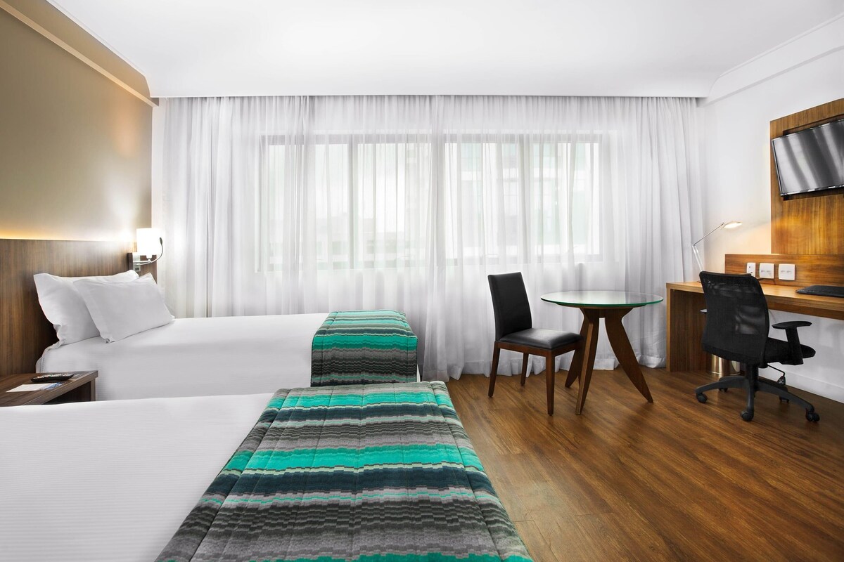 Hotellihuone, jossa on kaksi sänkyä, työpöytä, tuoli, televisio ja ikkuna, jossa on verhot.