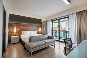 A hotel room with a bed, a sofa, a desk, and a balcony with a view of the city.