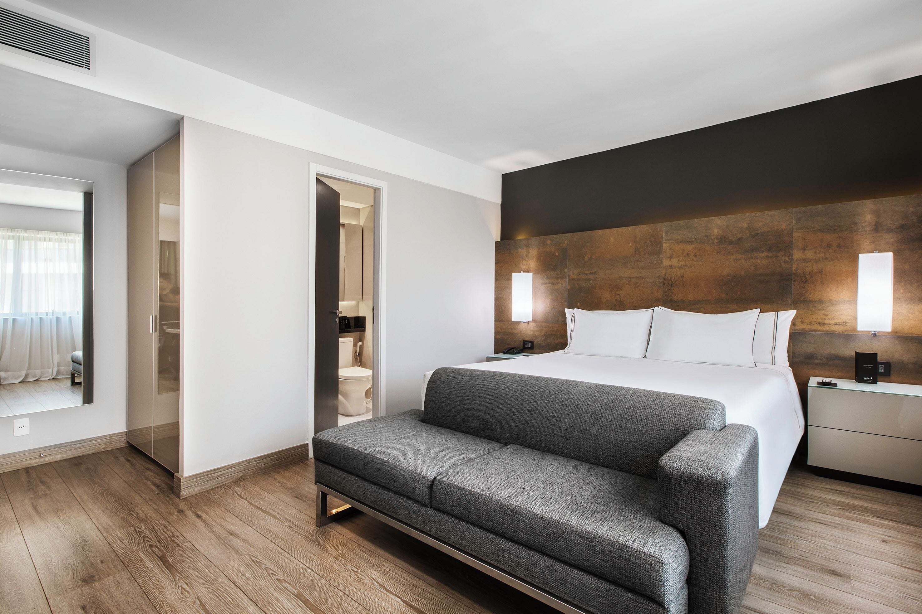 Et moderne hotelværelse med en stor seng, en sofa, trægulv, badeværelse med toilet og håndvask samt et stort spejl.