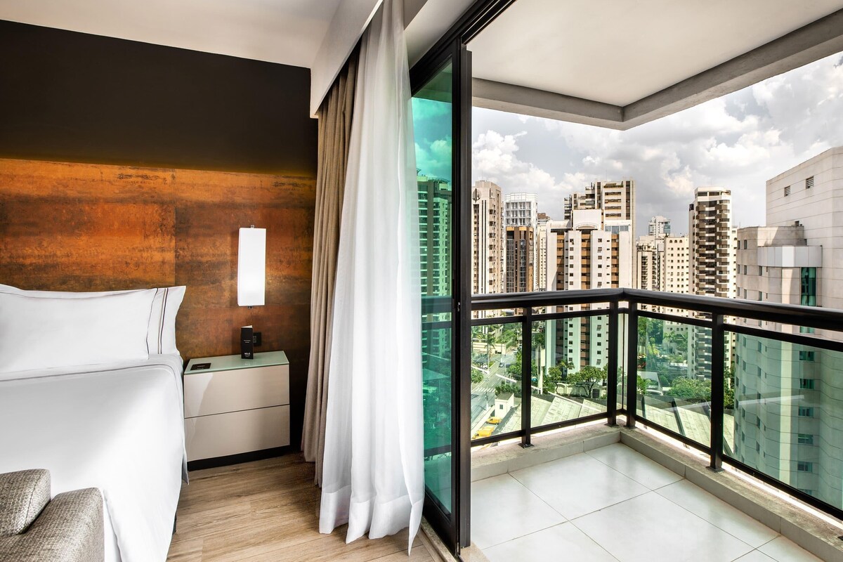 Una habitación de hotel moderna con balcón, cama, mesita de noche y vista a la ciudad.