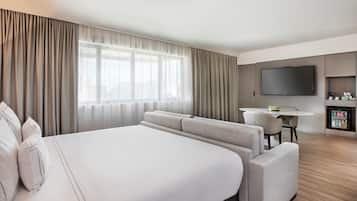 A hotel room with a large bed, a sofa, a dining table, a TV, and a minibar.