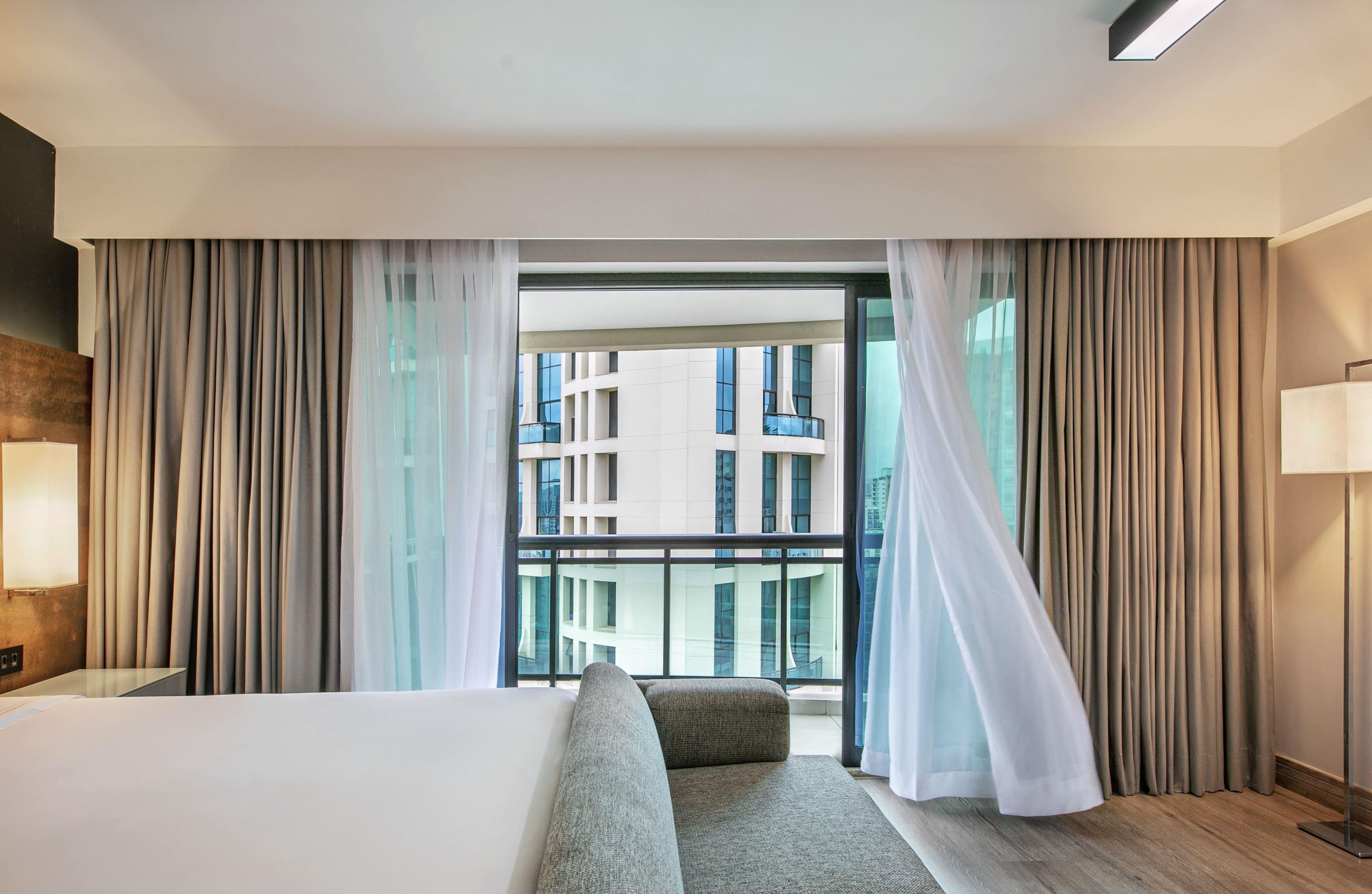 A hotel room with a large bed, a sofa, a view of a building through a window, and curtains.
