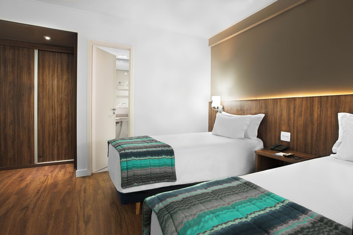 Una habitación de hotel con dos camas, suelo de madera, un armario y una mesita de noche con lámpara.