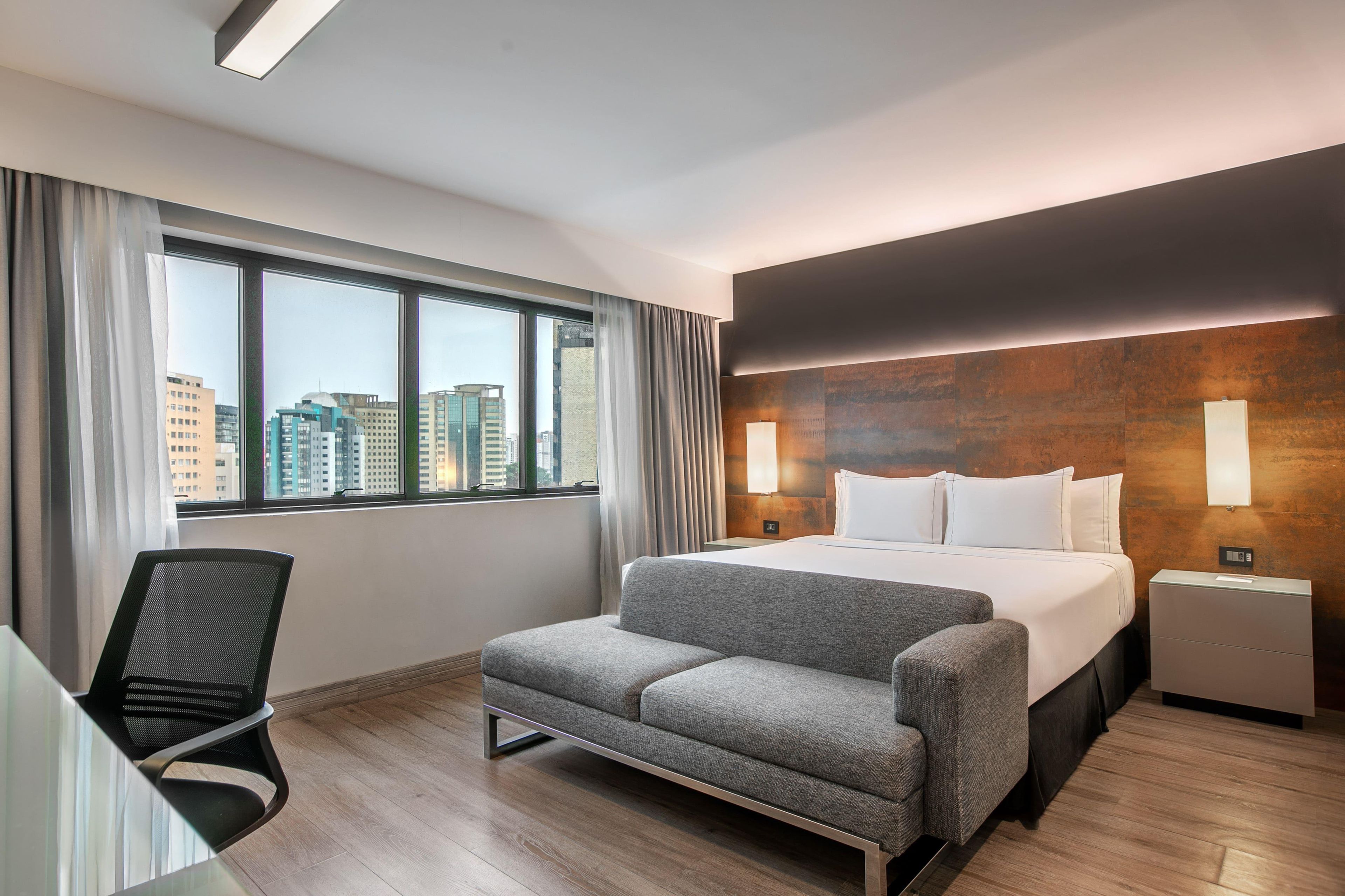 A modern hotel room with a bed, a grey sofa, a desk with a chair, and a large window offering a city view.