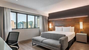 A modern hotel room with a bed, a grey sofa, a desk with a chair, and a large window offering a city view.